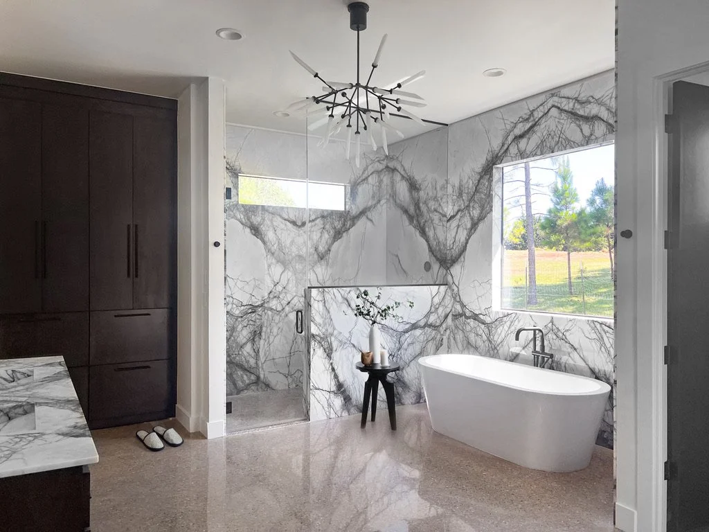 Freestanding bathtub beside a large window, marble-look slab walls, glass shower enclosure, dark wood cabinetry, terrazzo flooring, and modern pendant lighting.