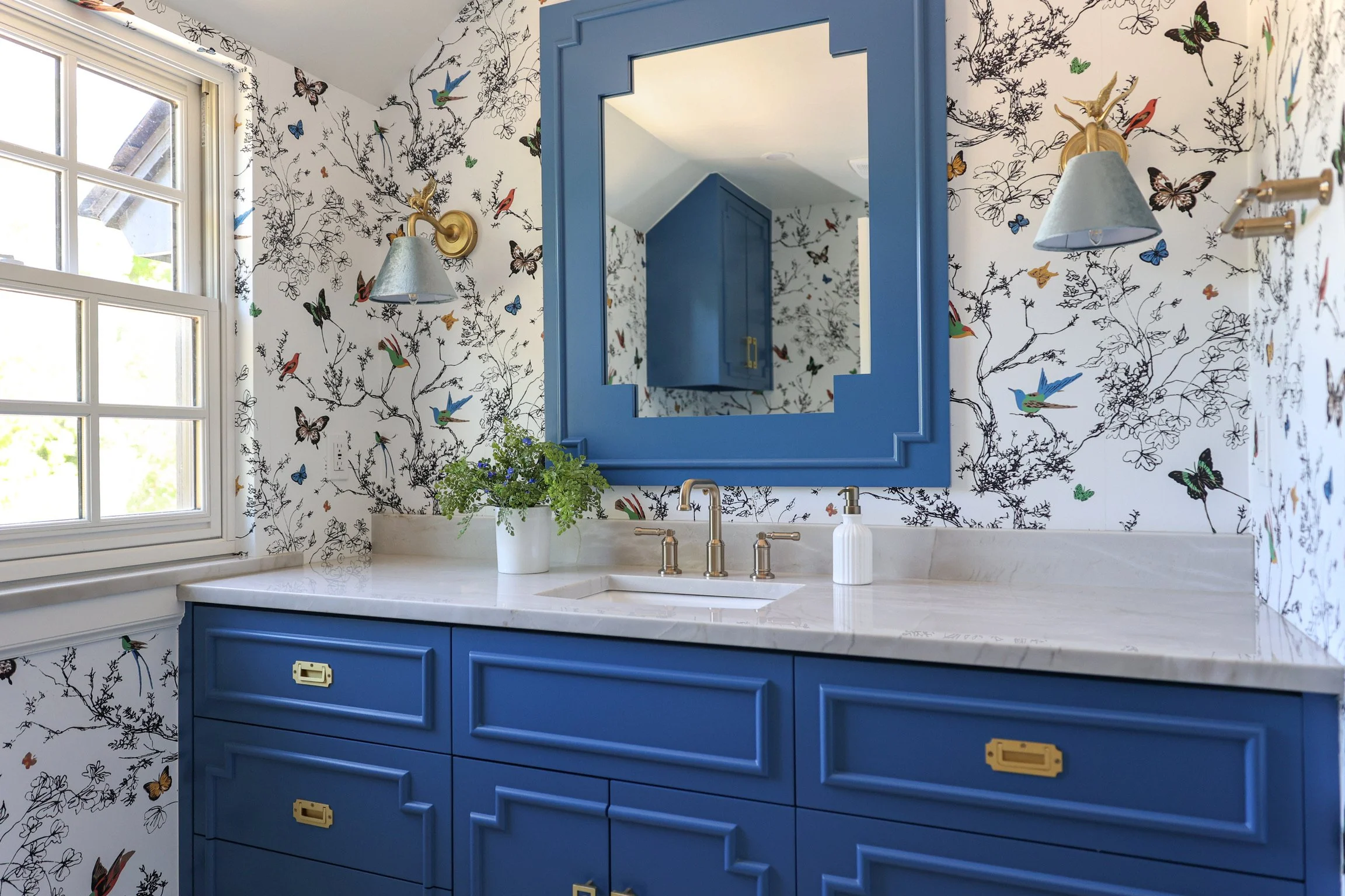 Blue bathroom vanity with brass hardware, marble countertop, framed mirror, brass sconces, and botanical wallpaper featuring birds and butterflies. Natural light from adjacent window.