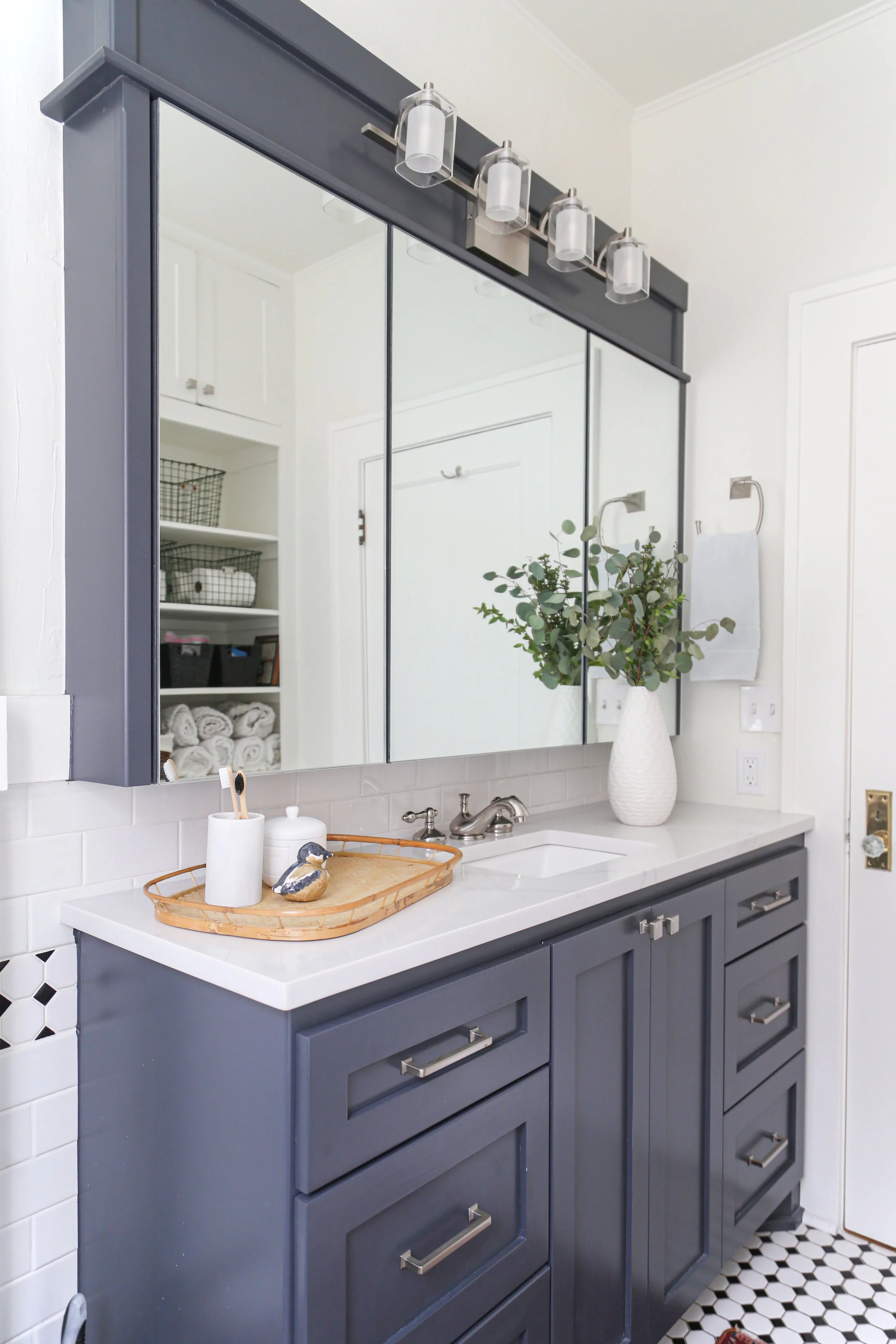 After renovation: Blue shaker-style vanity with quartz countertop, integrated sink, tri-panel mirrored medicine cabinet, nickel hardware, white subway tile backsplash, hexagon floor tile, and built-in linen storage.