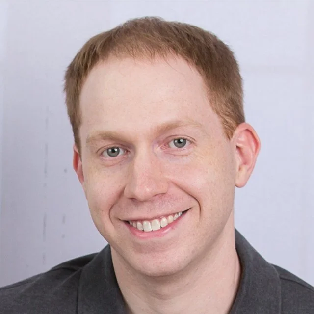 Professional headshot of a smiling man with short light brown hair, wearing a dark collared shirt against a neutral background, representing a member of the research team.