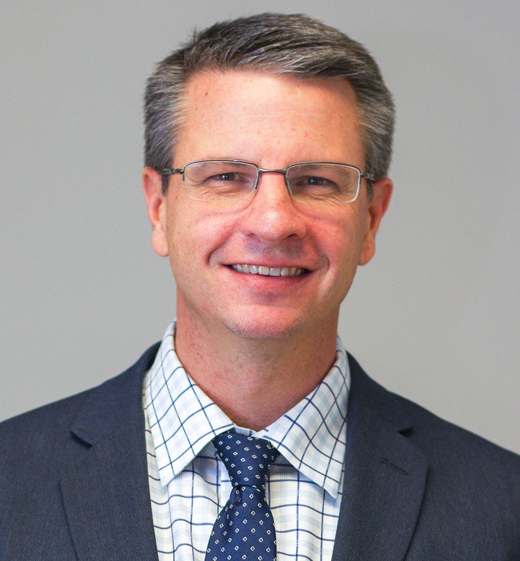 Professional headshot of a smiling man with short gray hair and glasses, wearing a suit jacket, patterned shirt, and tie against a neutral background, representing a senior member of the research team.