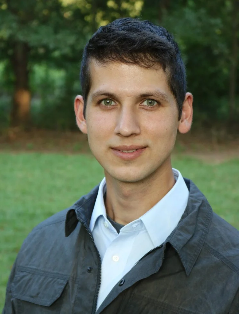 Professional headshot of a man with short dark hair and green eyes, wearing a collared shirt and jacket, photographed outdoors with a blurred green background, representing a member of the research team.