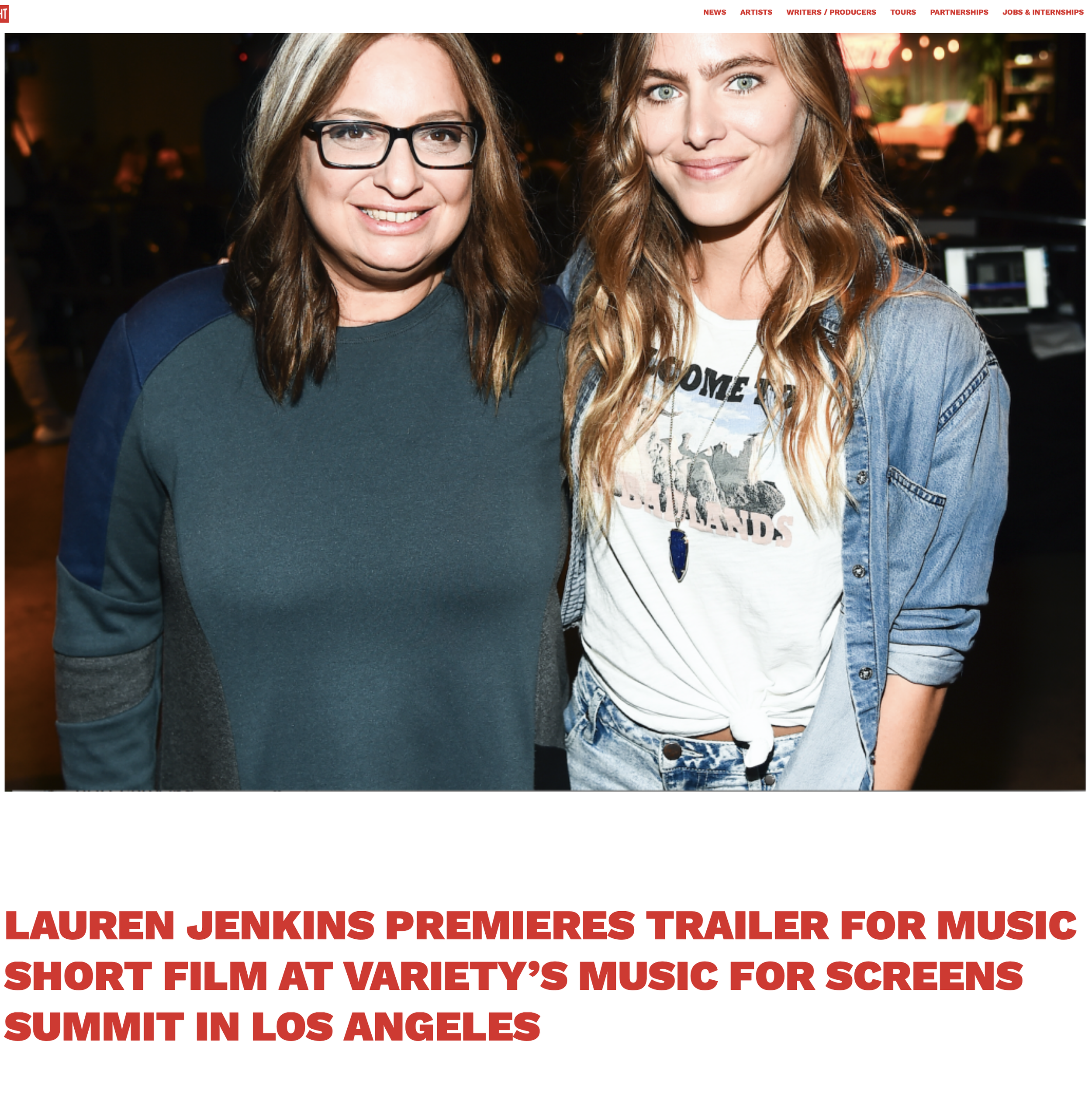 Two women smiling, Lauren Jenkins at a music event in Los Angeles, one wearing glasses and a green and blue sweatshirt, the other with long wavy hair, a white graphic t-shirt, and a denim jacket.