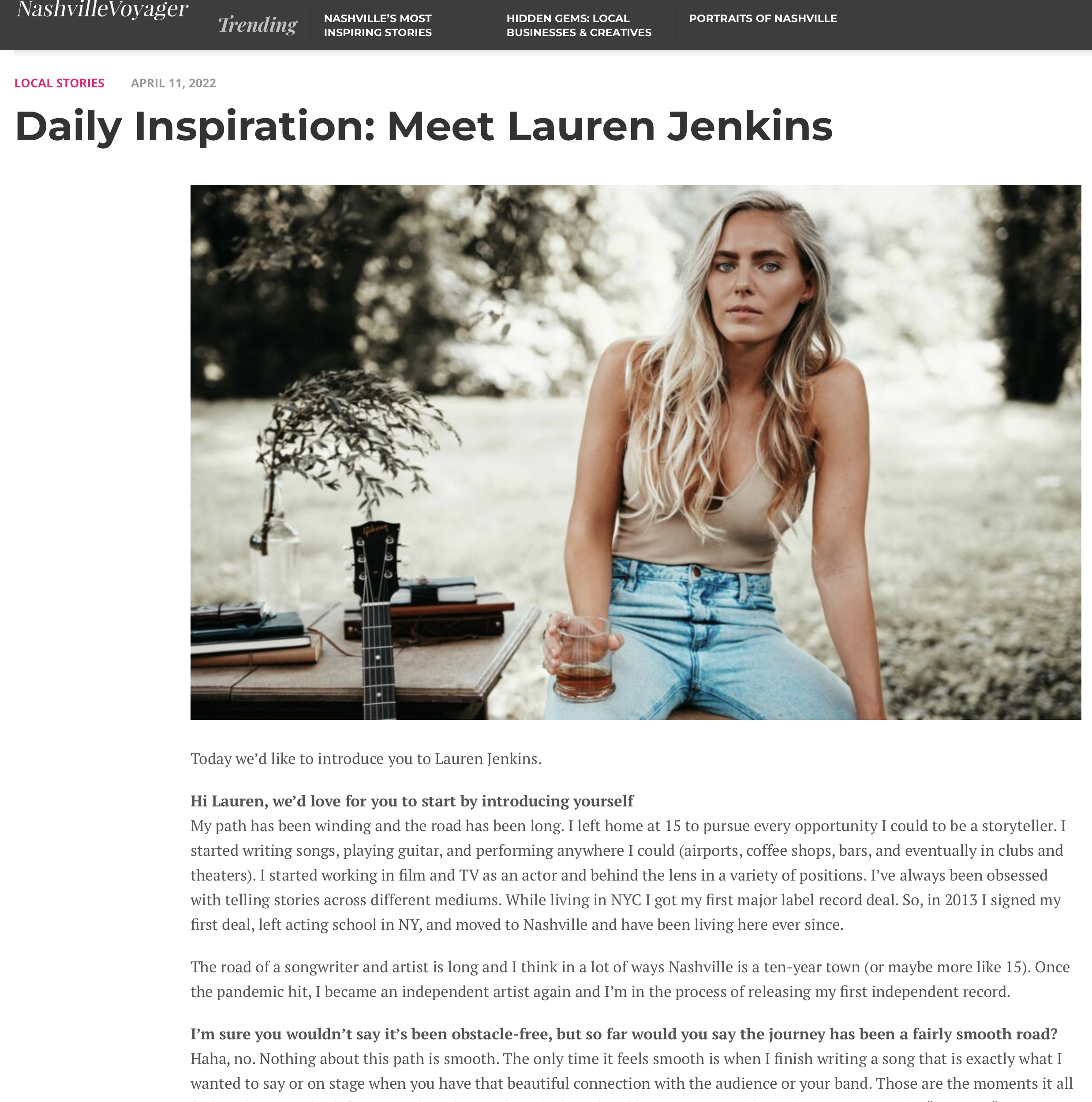Lauren Jenkins with long blonde hair, wearing a beige tank top and denim jeans, sitting outdoors in a park-like setting, holding a glass of drink. There is a small table with a potted plant, some books, and a guitar beside her.
