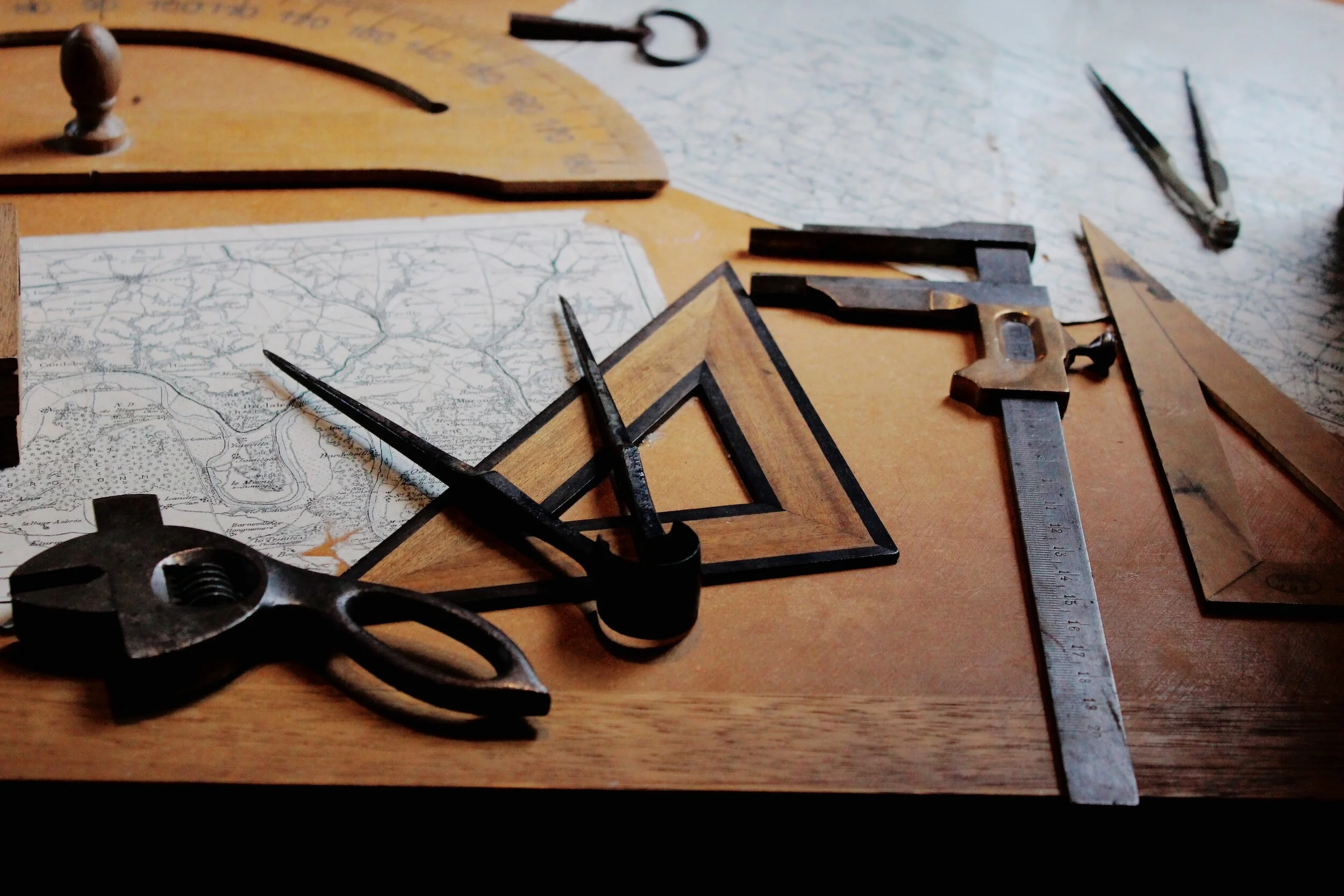 Old drafting tools and maps on a wooden table, including calipers, square, compass, and protractor.
