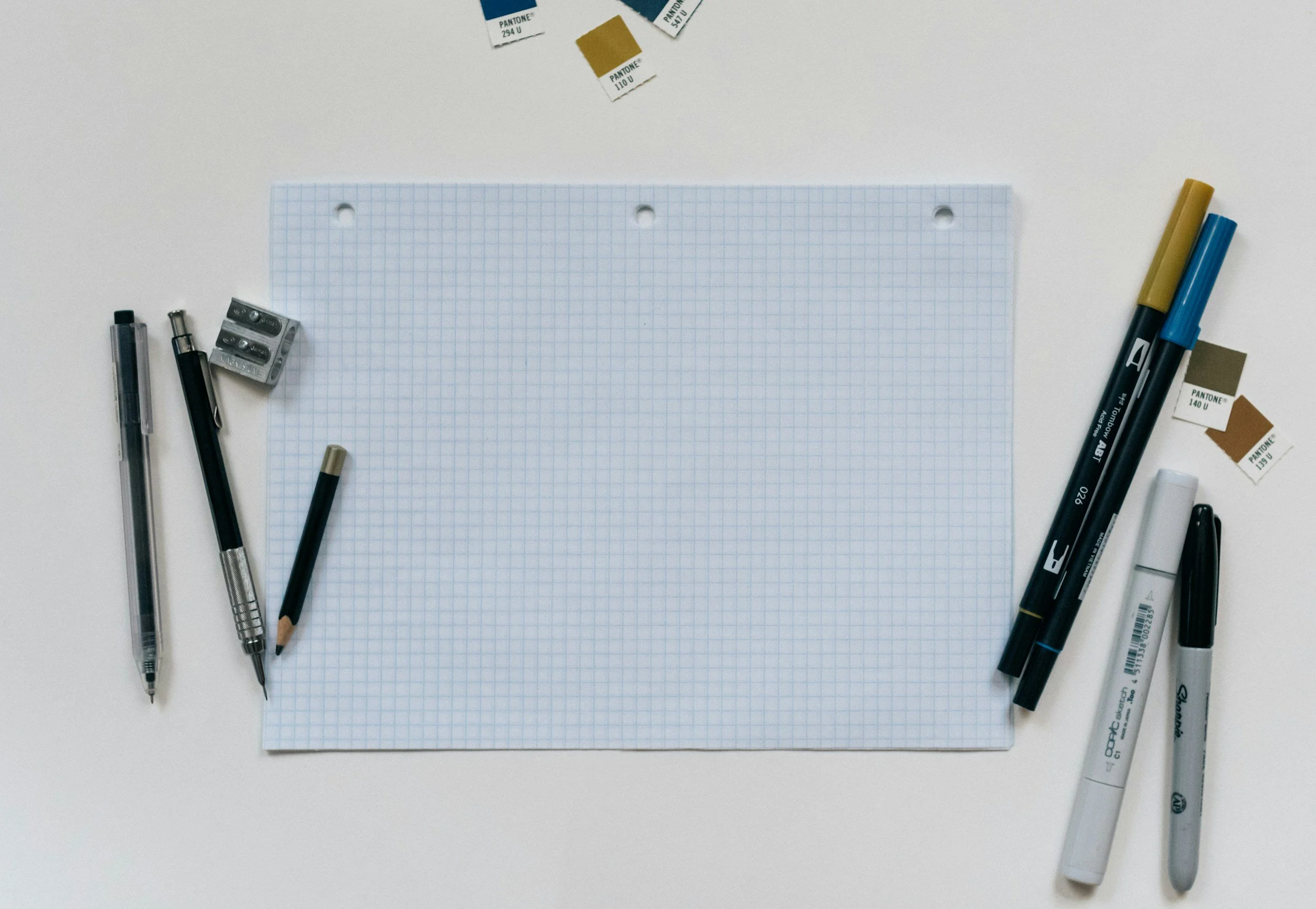 A sheet of graph paper on a white surface surrounded by various stationery items including pens, pencils, a pencil sharpener, markers, and Pantone color swatches.