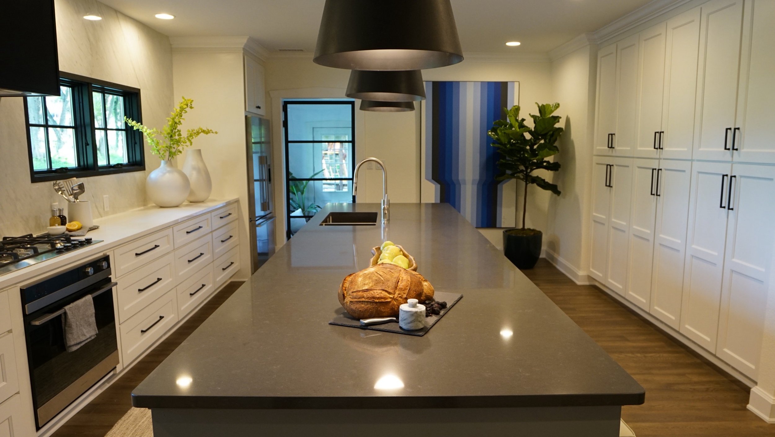 Modern kitchen with long island, dark countertops, pendant lights, potted plant, decorative vases, bread and fruit on counter, and white cabinetry.