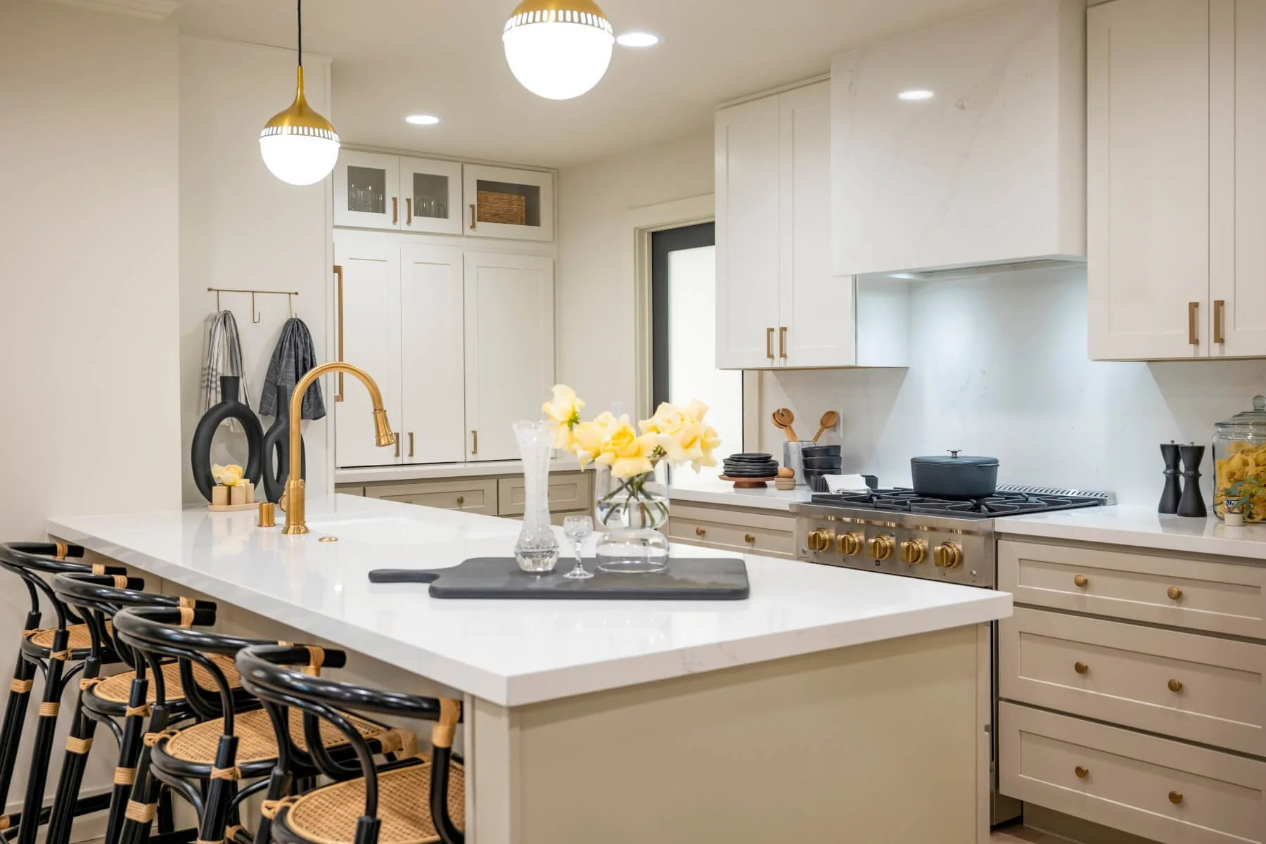 Modern kitchen with white cabinets, brass fixtures, and a large island featuring bar stools. The countertop has a vase with yellow roses and decorative glassware. The stove features gold-colored knobs, and there are pendant lights overhead.