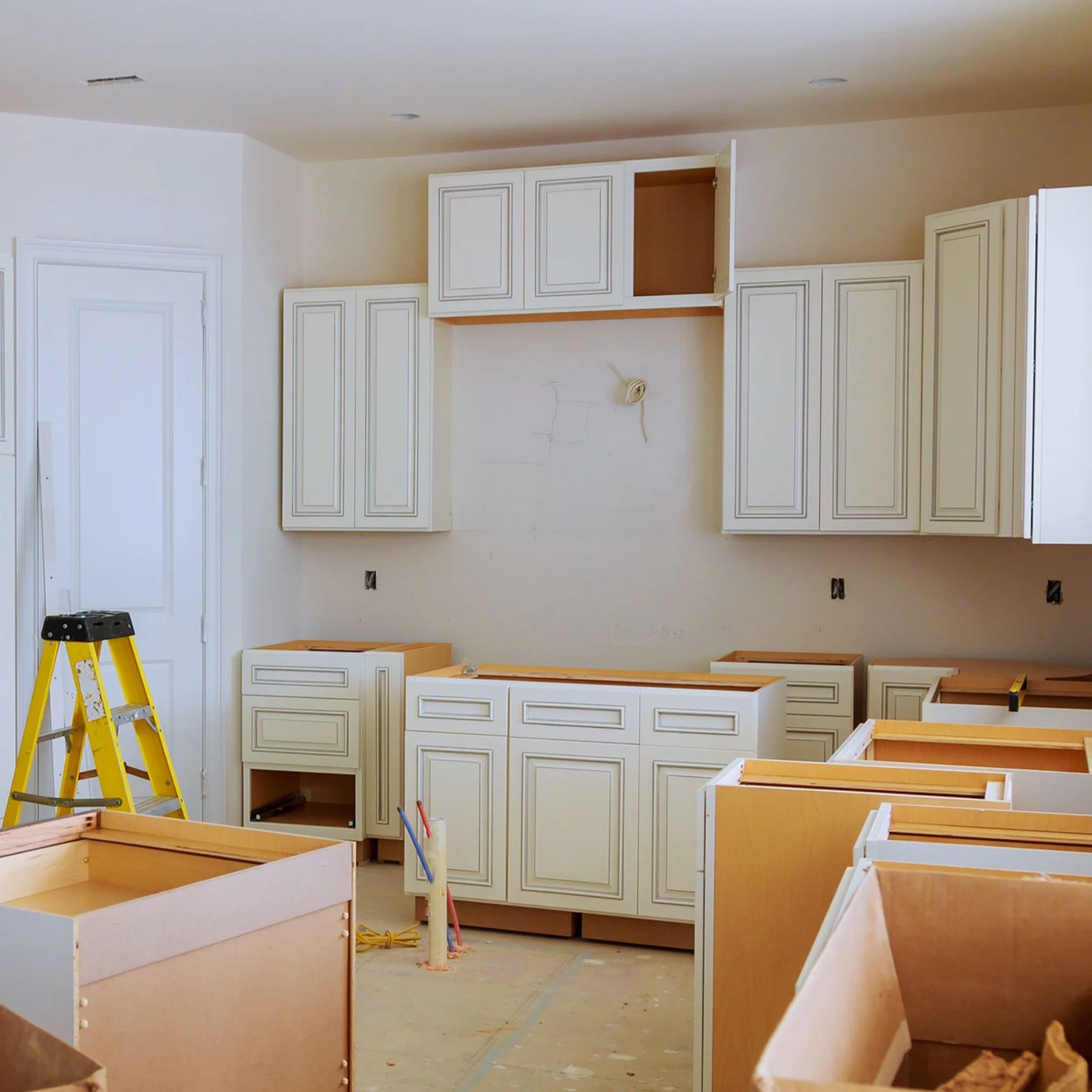 A partially installed kitchen with white cabinetry, unpainted walls, and a yellow step ladder. Some cabinets have doors open or missing, and construction tools are present.