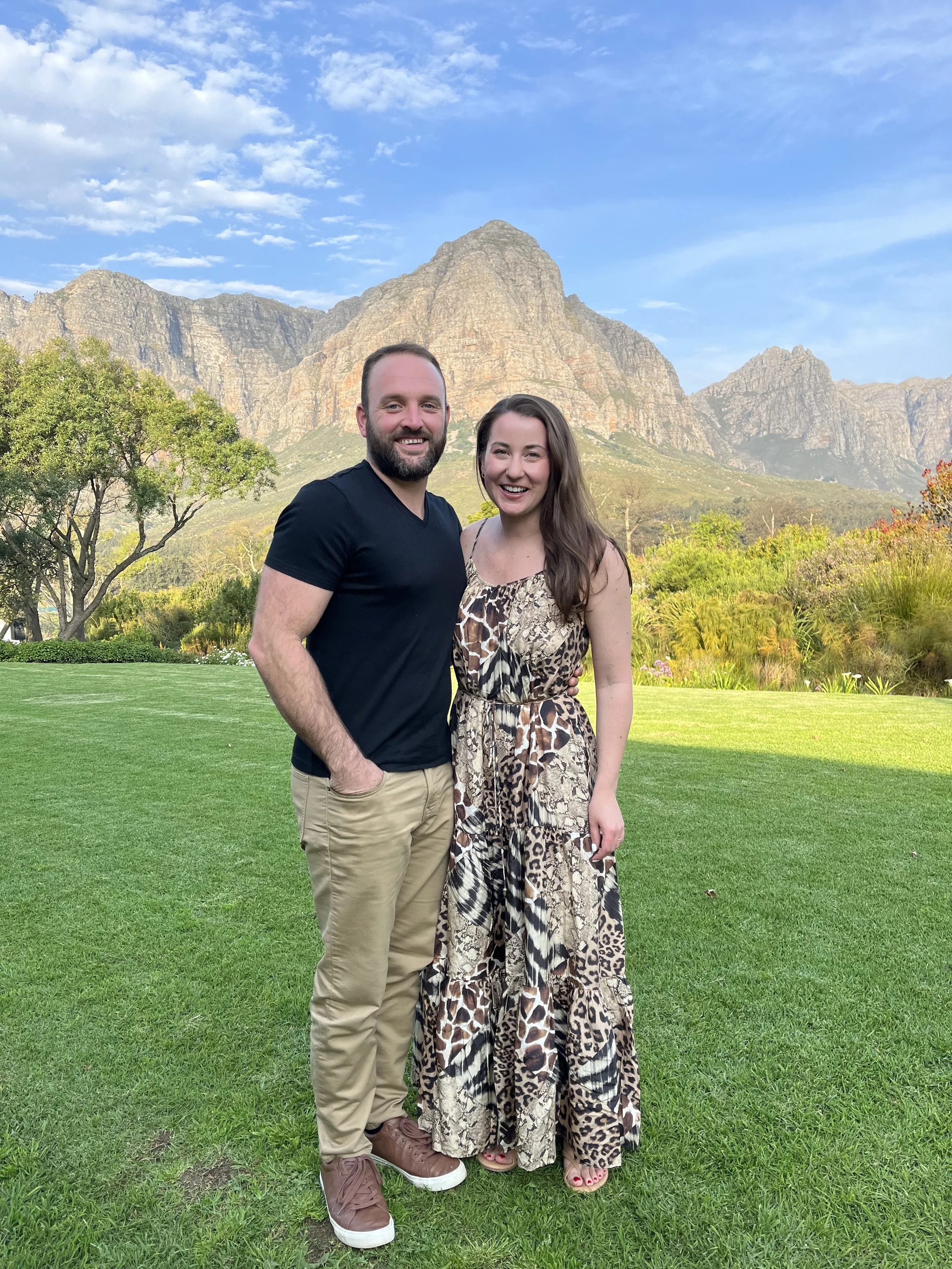 A man and a woman standing together on a grassy area with mountain scenery in the background.