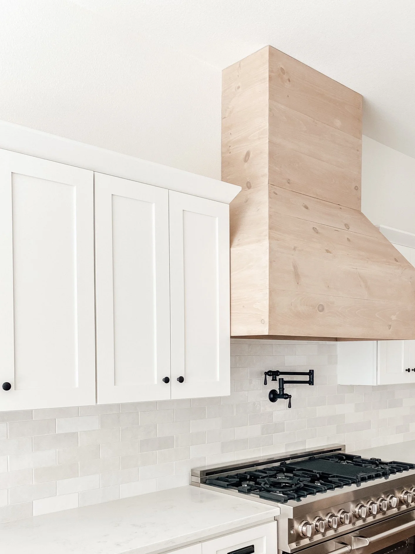 Modern kitchen with white cabinets, a light wood range hood, stainless steel stove, and a pot filler above the stove. The backsplash features light-colored subway tiles.