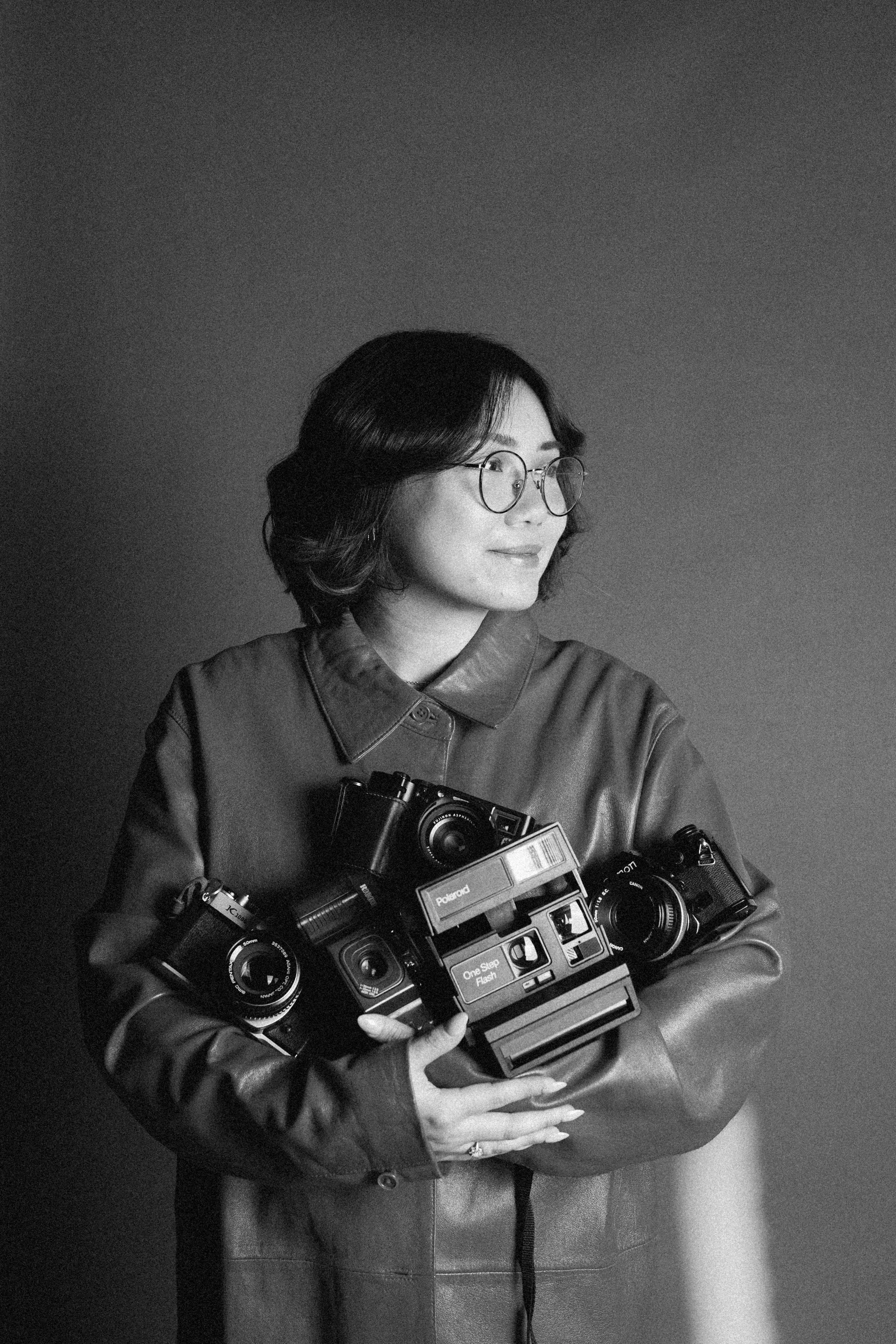 A woman with short dark hair, glasses, and a leather jacket holding multiple vintage cameras, standing in front of a plain, neutral background.
