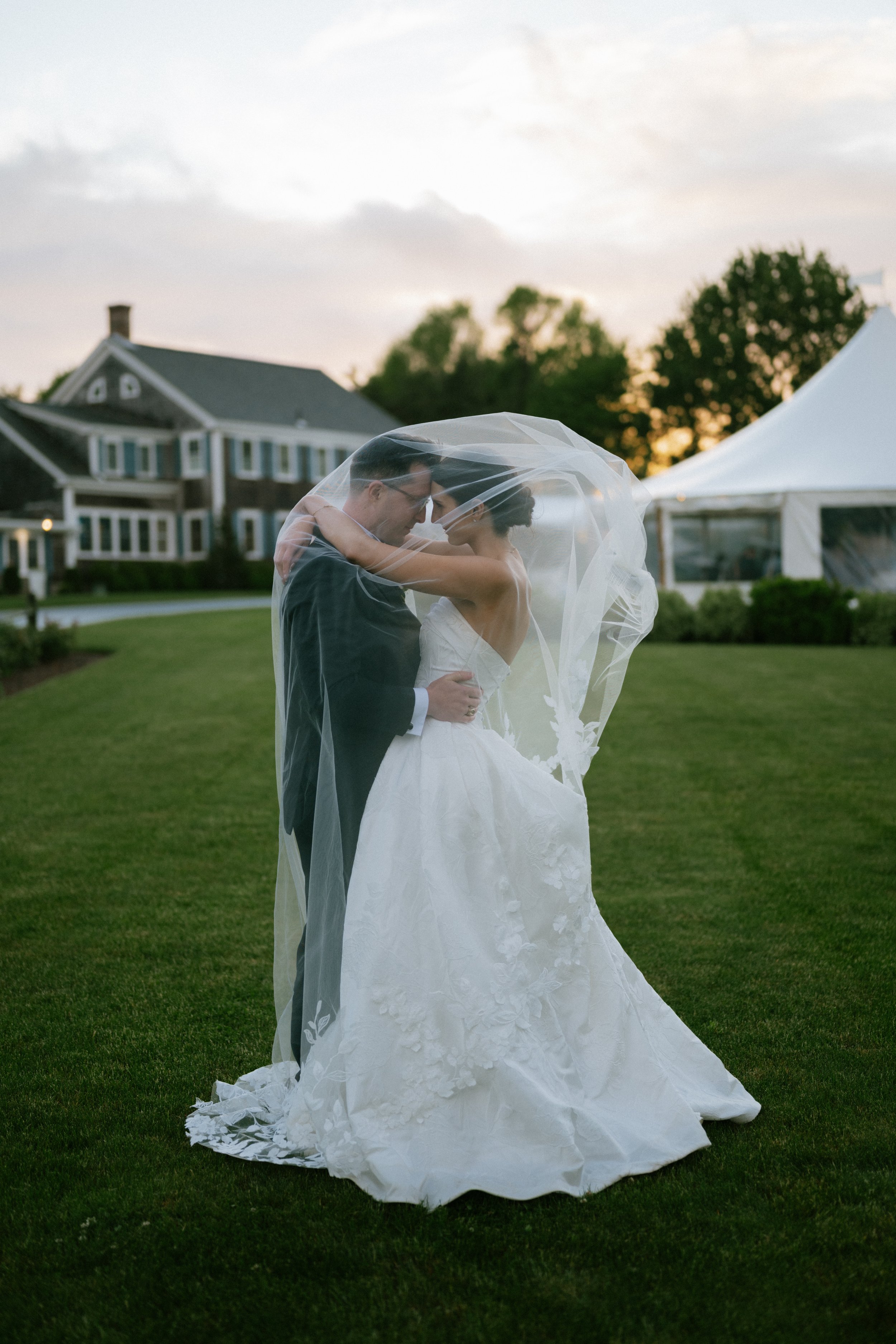 A bride and groom sharing a dance under a veil on a grassy area at sunset, with a house and trees in the background.