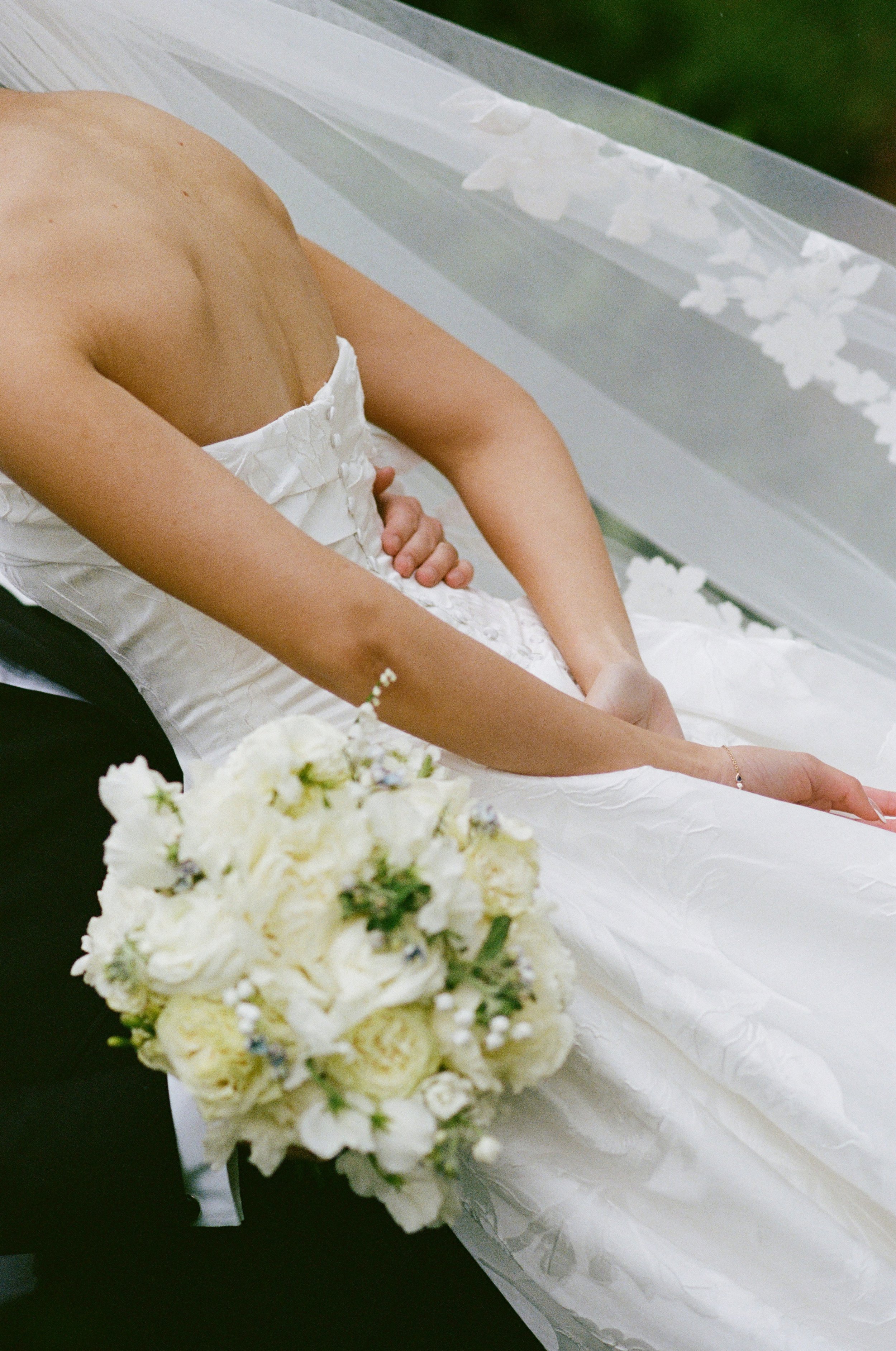 A bride in a strapless white wedding gown holding a bouquet of white flowers, standing near a window with floral curtain.