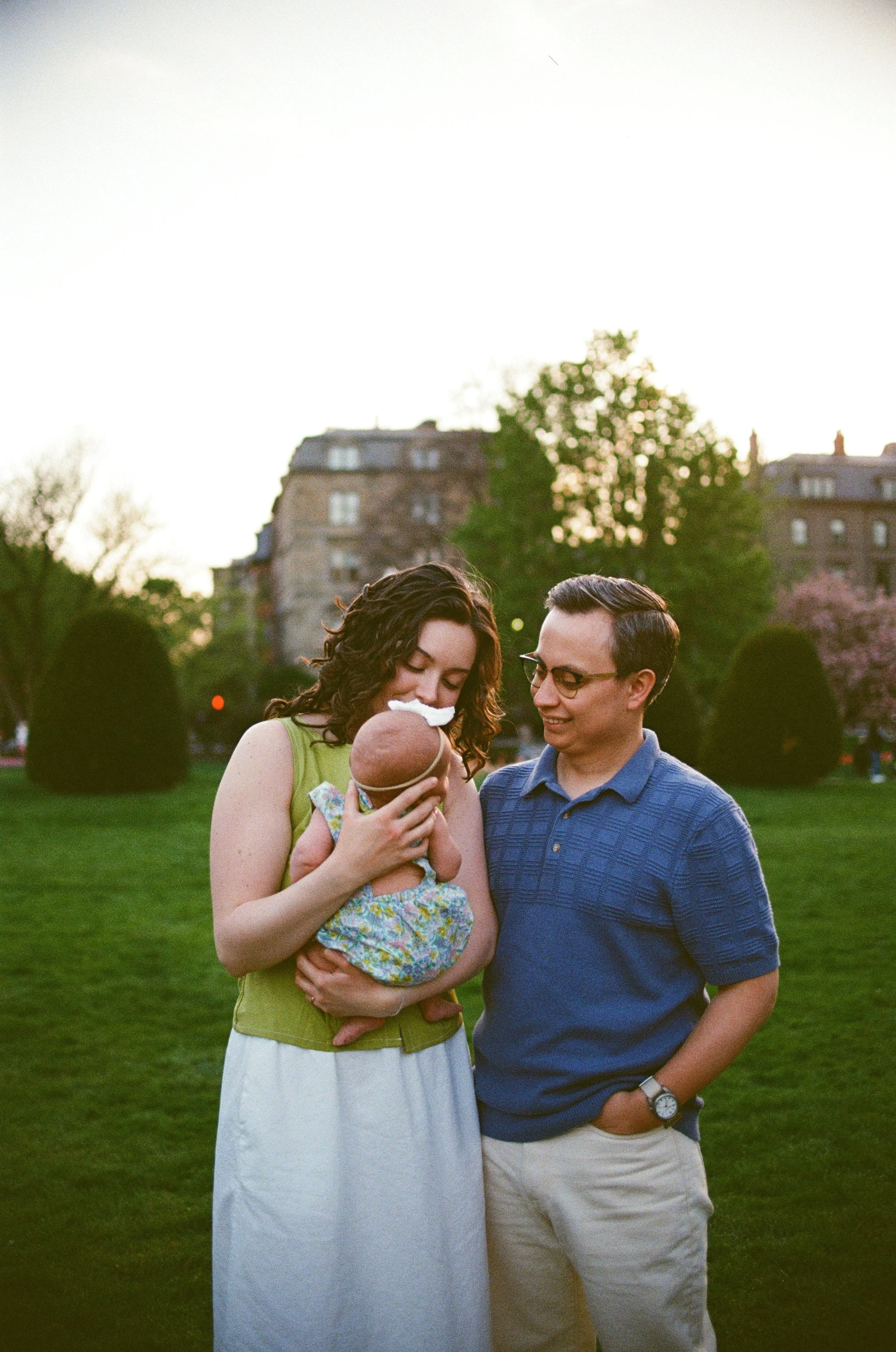 A family outside in a park during sunset. The mother is holding a newborn baby, and the father is standing beside her, smiling and looking at the baby. Trees and buildings are visible in the background.