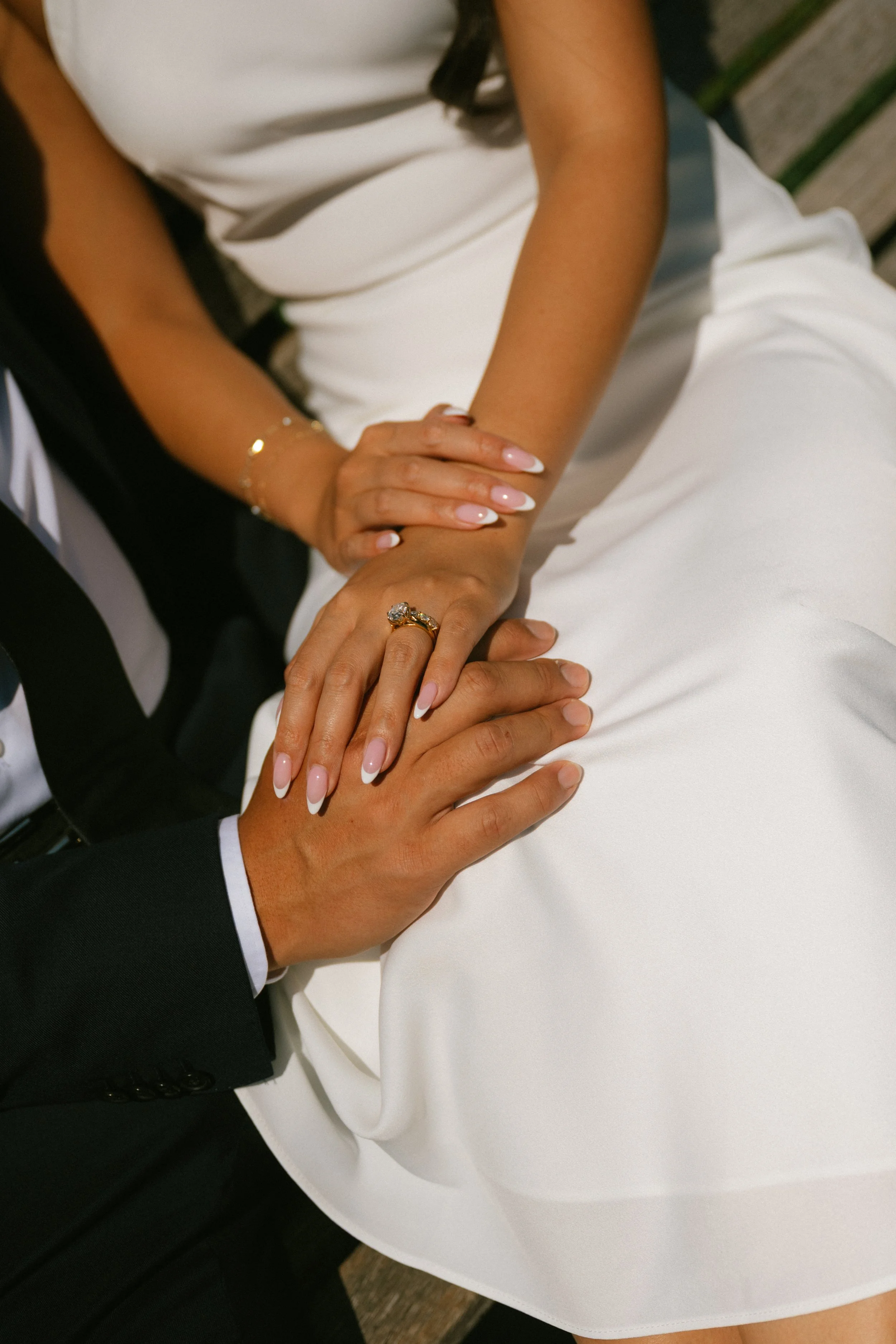 Close-up of a couple holding hands, showing wedding rings, with a woman wearing a white dress and a man in a black suit.