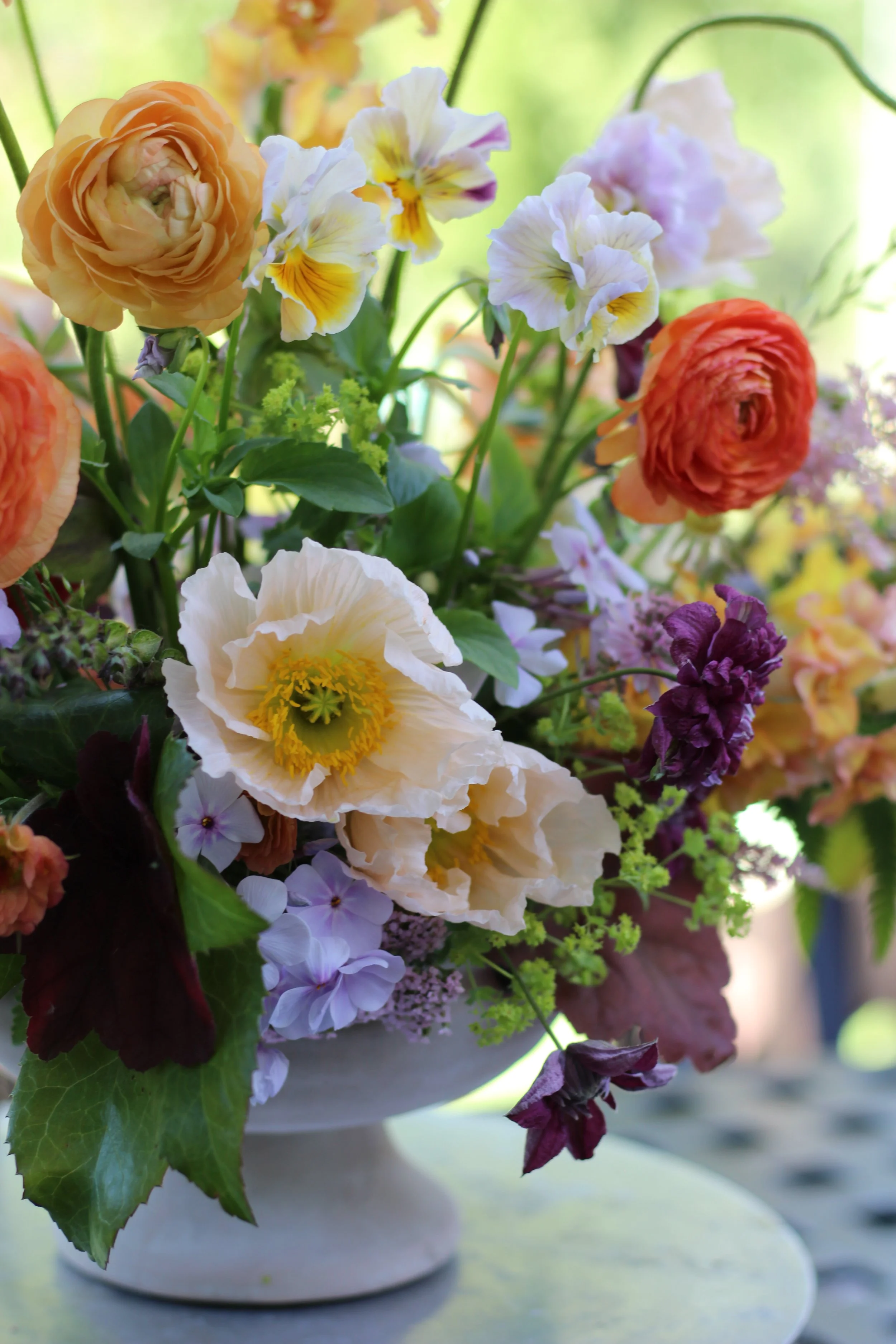 A close-up of a colorful flower arrangement in a white vase, featuring orange, white, purple, and yellow flowers with green leaves, set on a table with a blurred outdoor background.