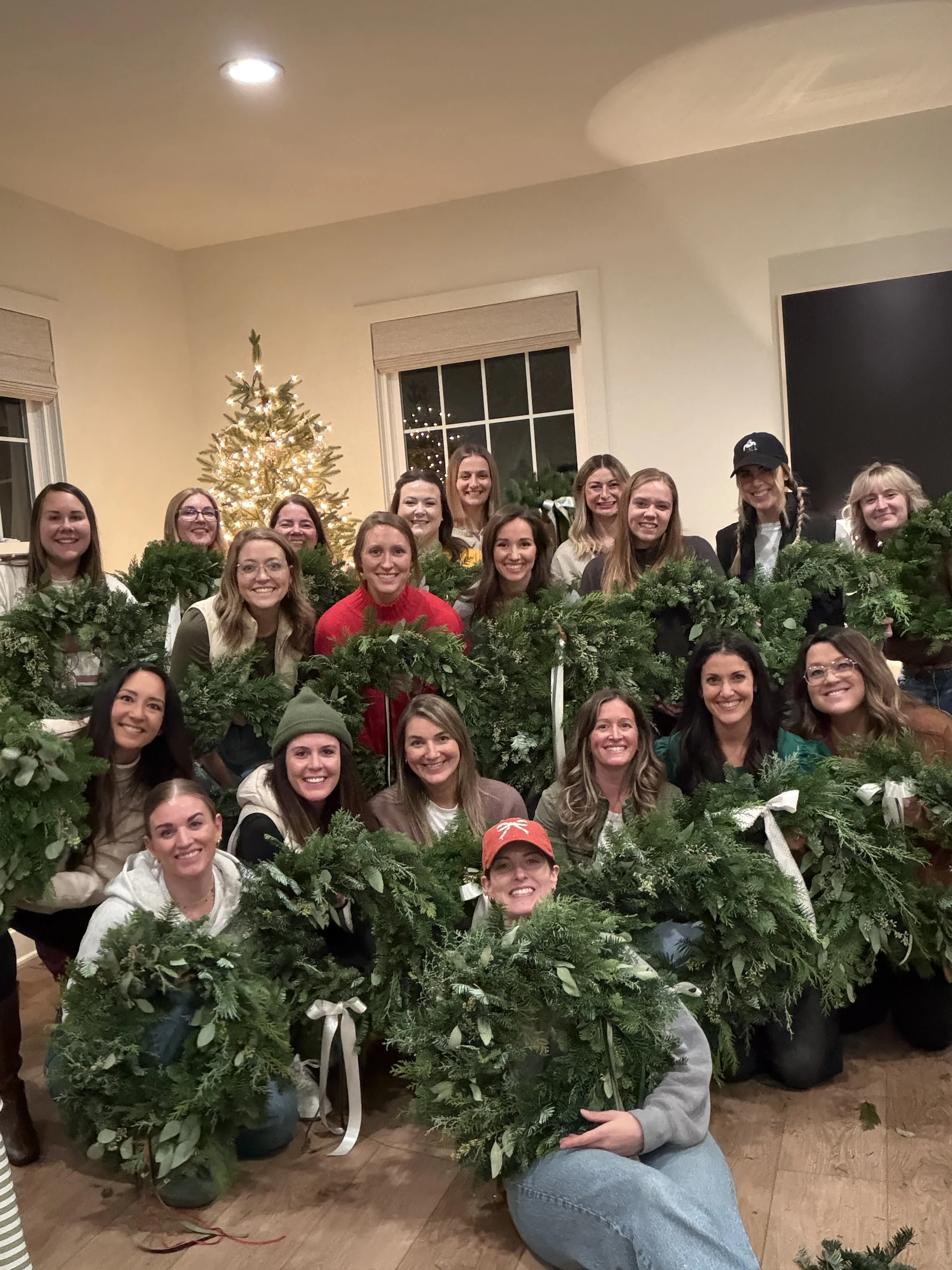 Group of women holding Christmas wreaths in a festive room with a decorated Christmas tree and windows in the background.