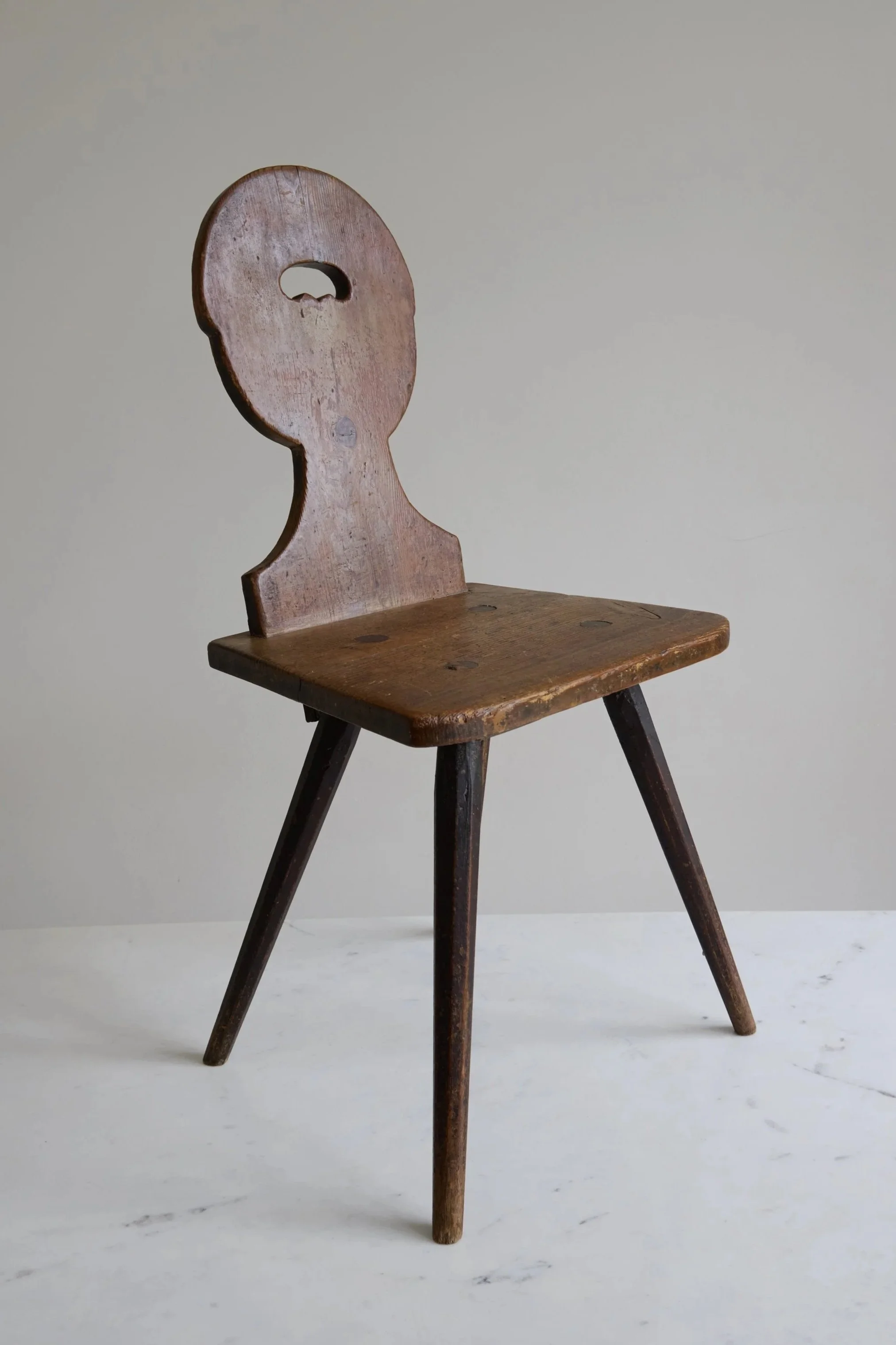 An old wooden chair with a backrest shaped like a face, sitting on a white surface against a plain gray background.