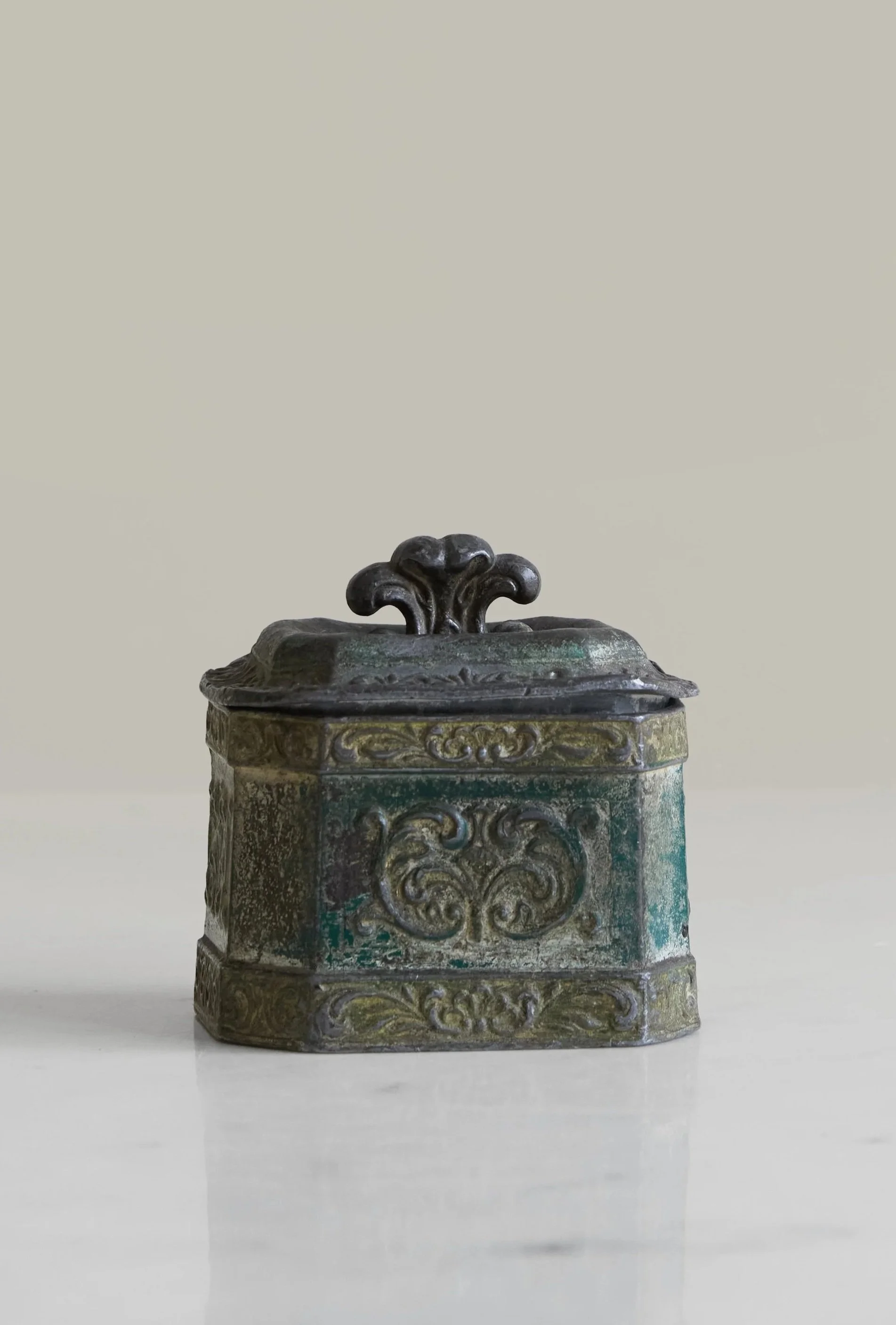 An ornate, antique metal box with intricate carvings and a dark decorative handle on the lid, resting on a smooth, white surface.