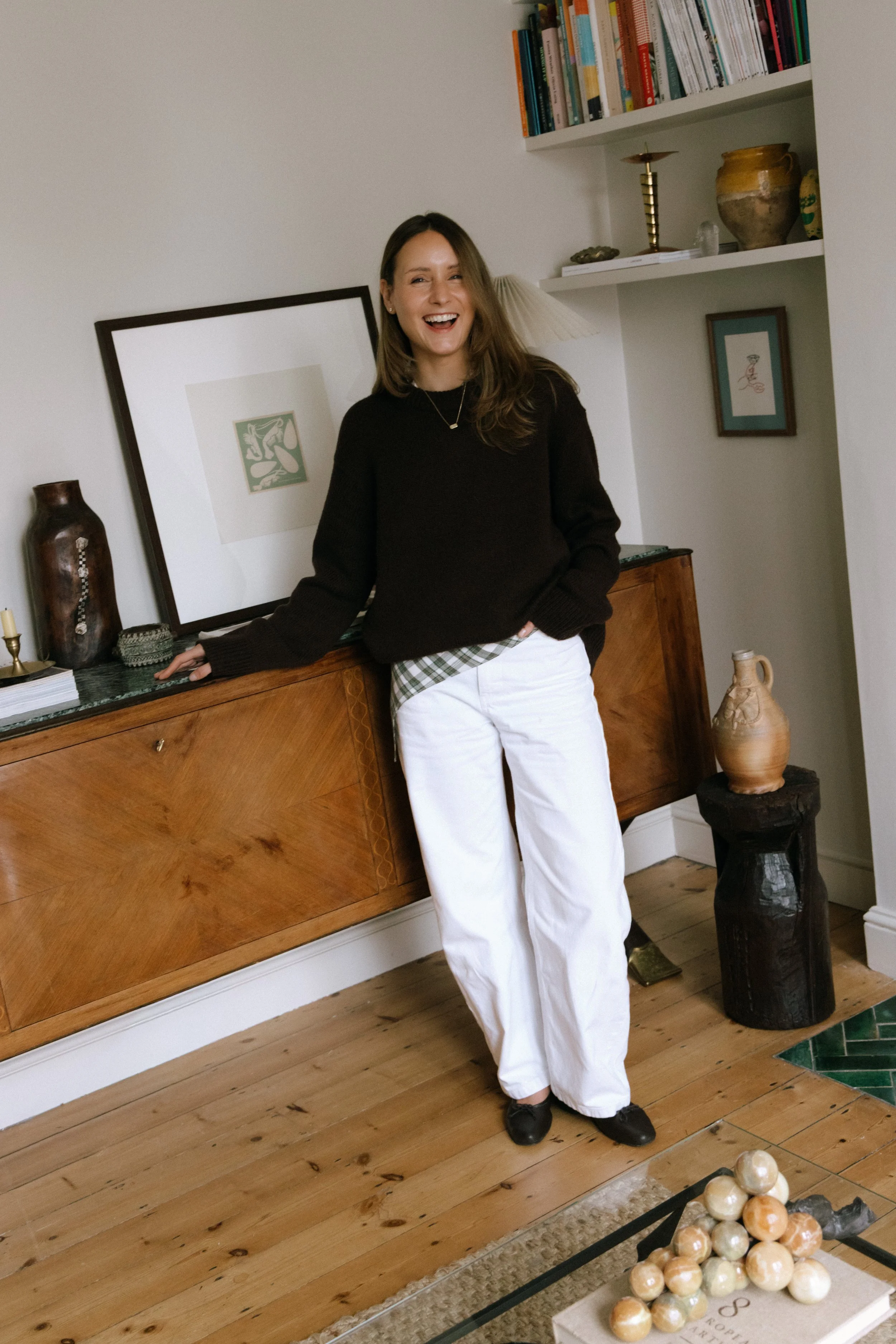 A woman with long brown hair laughing and leaning against a wooden sideboard in a living room. She is wearing a black sweater, white pants, and black shoes, with a green plaid shirt underneath. There are books, vases, and artwork on the wall behind her, along with a small sculpture of spheres on a coffee table in front of her.