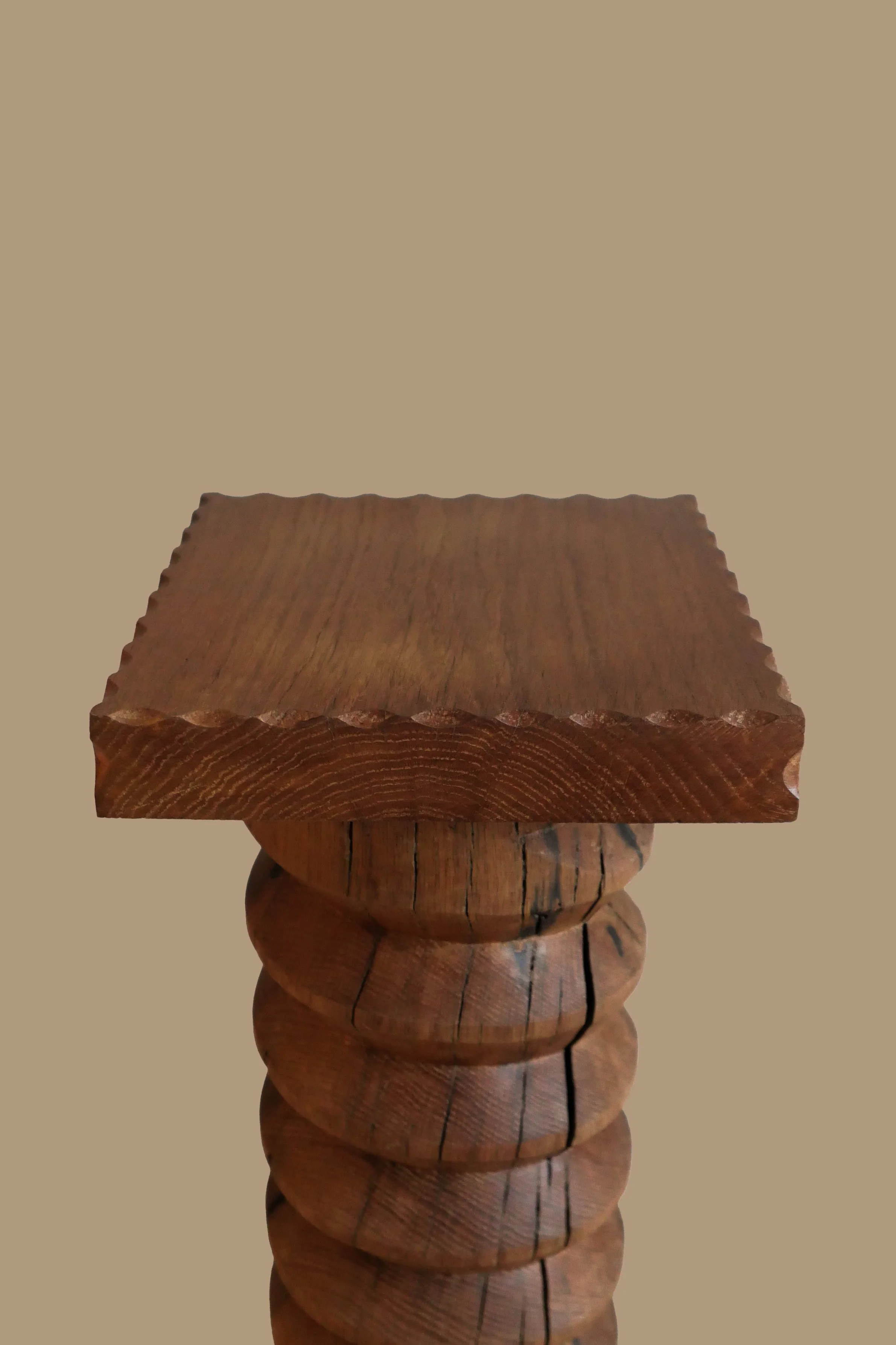Close-up of a wooden table with a square top and a carved, turned wood pedestal base against a plain beige background.