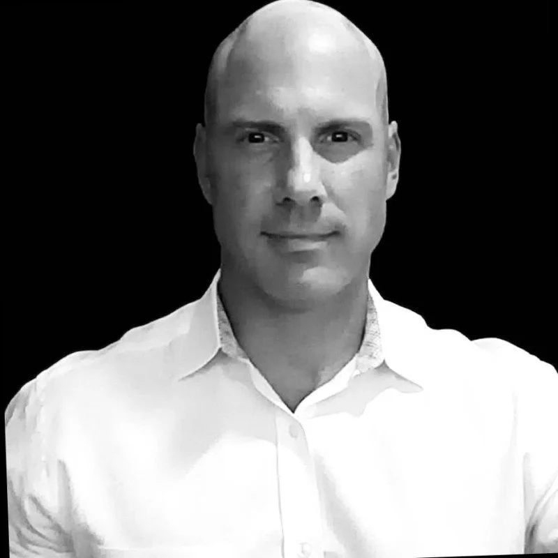 A black-and-white portrait of a bald man in a white collared shirt, smiling slightly, against a black background.