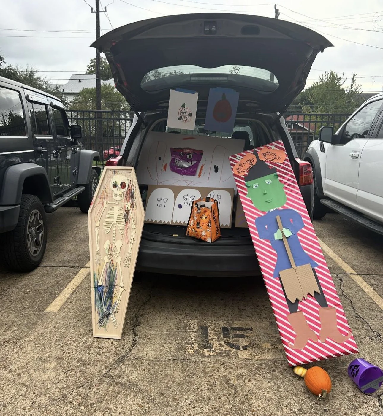 Halloween-themed decorations and artwork displayed in the back of a parked vehicle, including a skeleton drawing, a large Frankenstein figure, and various other Halloween illustrations on painted boards and hanging posters, with small pumpkins and Ha