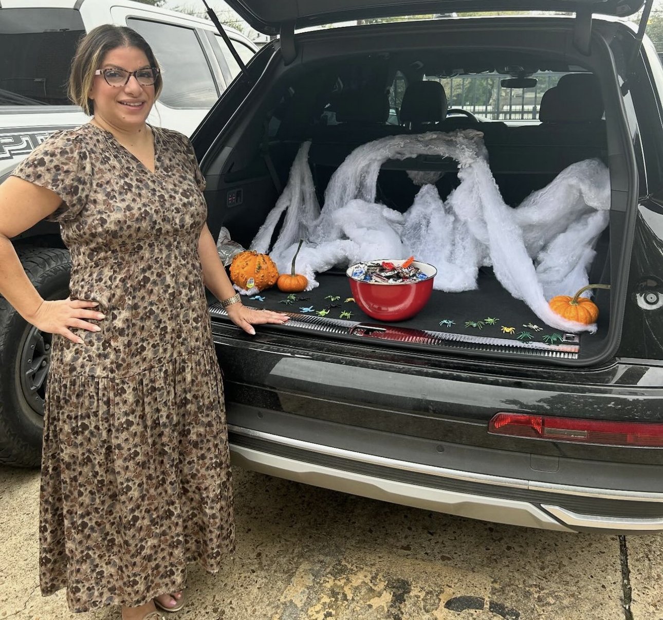 Woman standing next to a decorated car trunk with a Halloween-themed display, including fake spider webs, small pumpkins, and a bowl of Halloween candies.