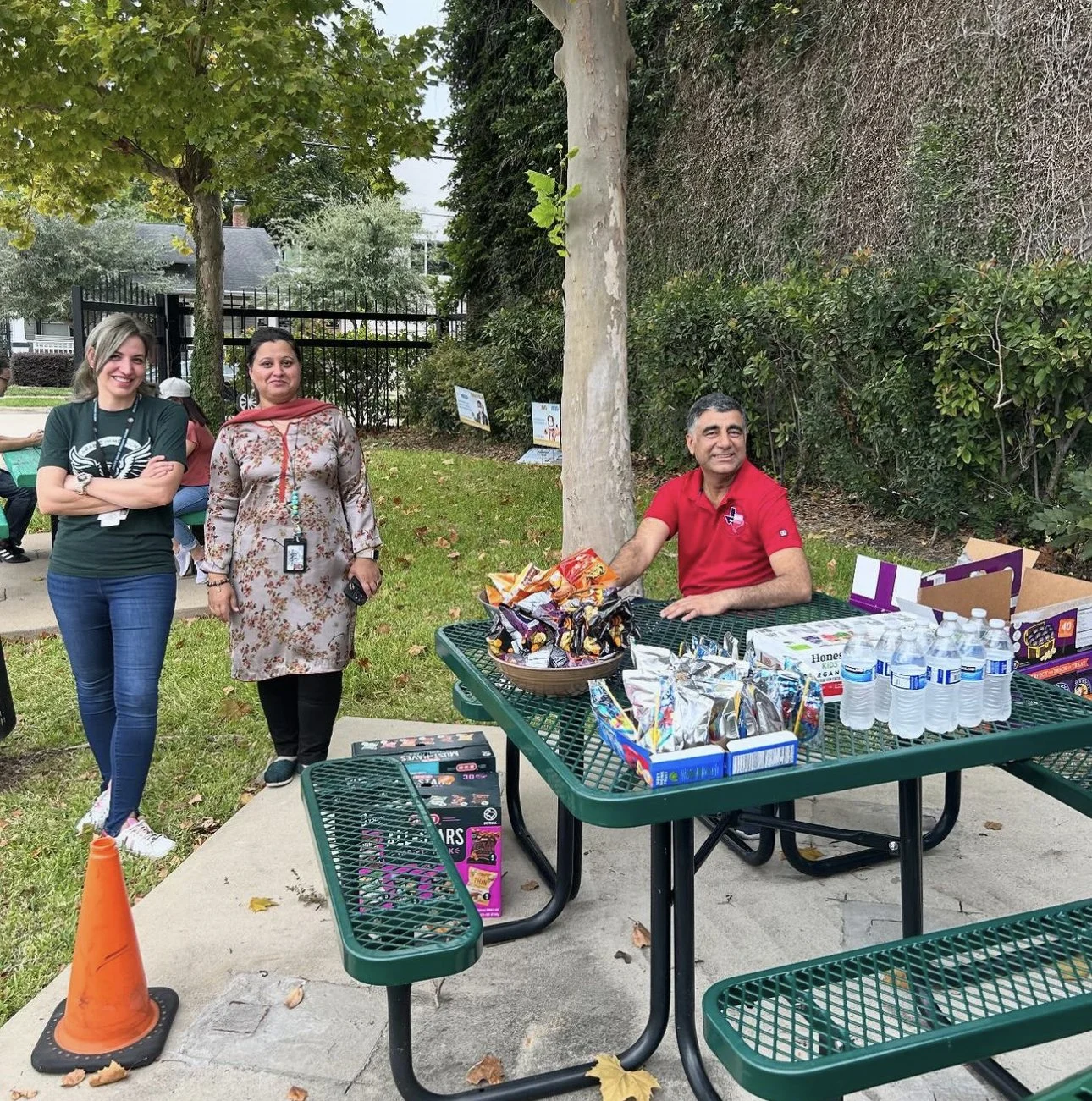 People at an outdoor event near a green picnic table, with snacks, bottled water, and a man in a red shirt sitting behind the table, smiling. Two women are standing nearby, one in a dark green shirt and the other in a floral dress.