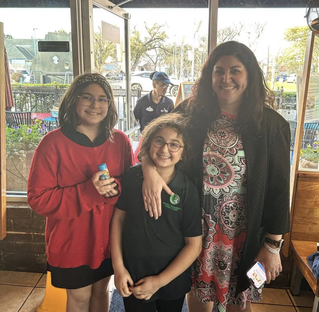 Three smiling girls and a woman standing inside a restaurant, with windows in the background showing an outdoor street scene. One girl holds a glue stick, and another girl wears glasses and a black shirt.