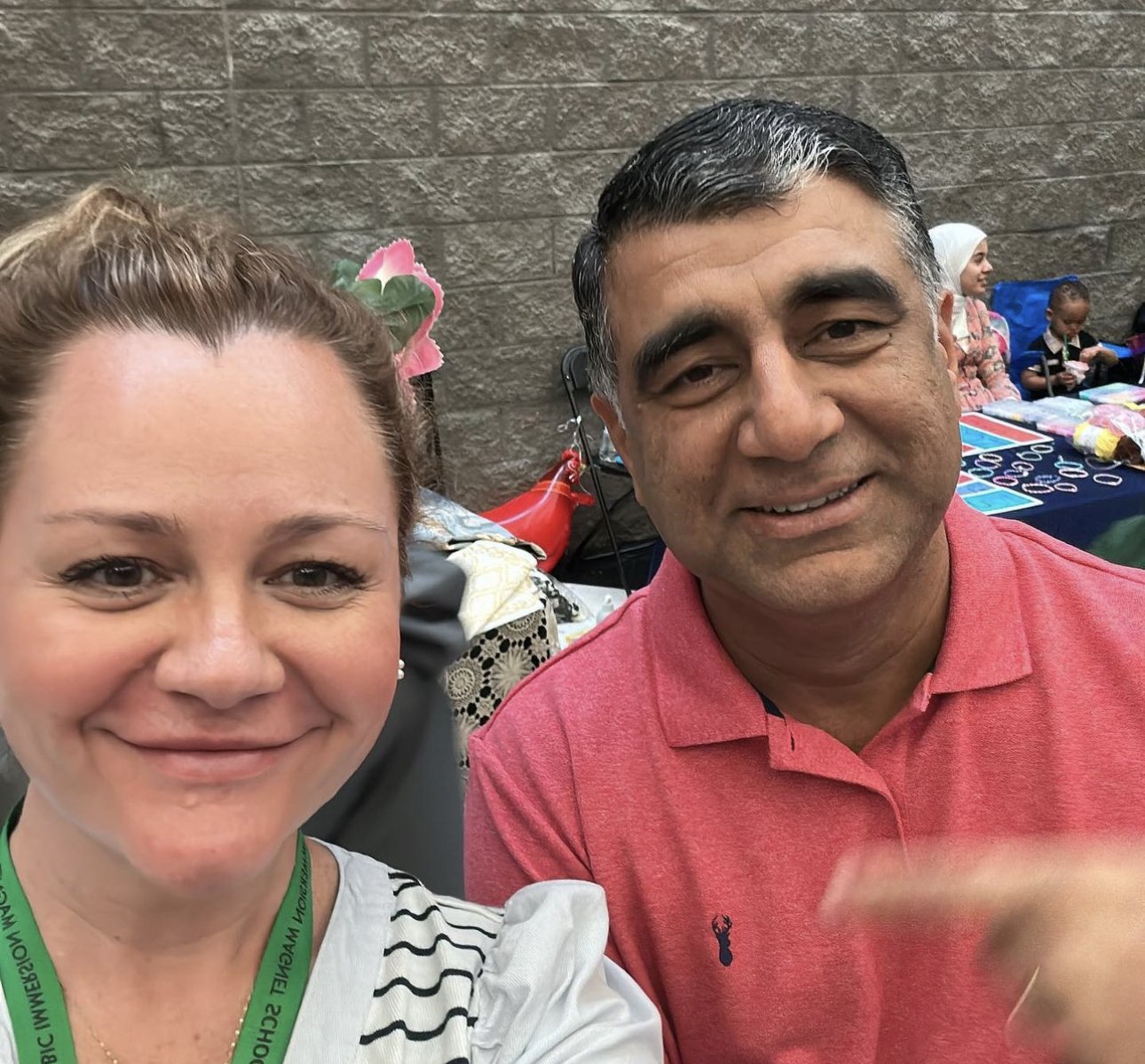 A woman and a man taking a selfie at an indoor event with other people in the background, some sitting at tables with colorful items.
