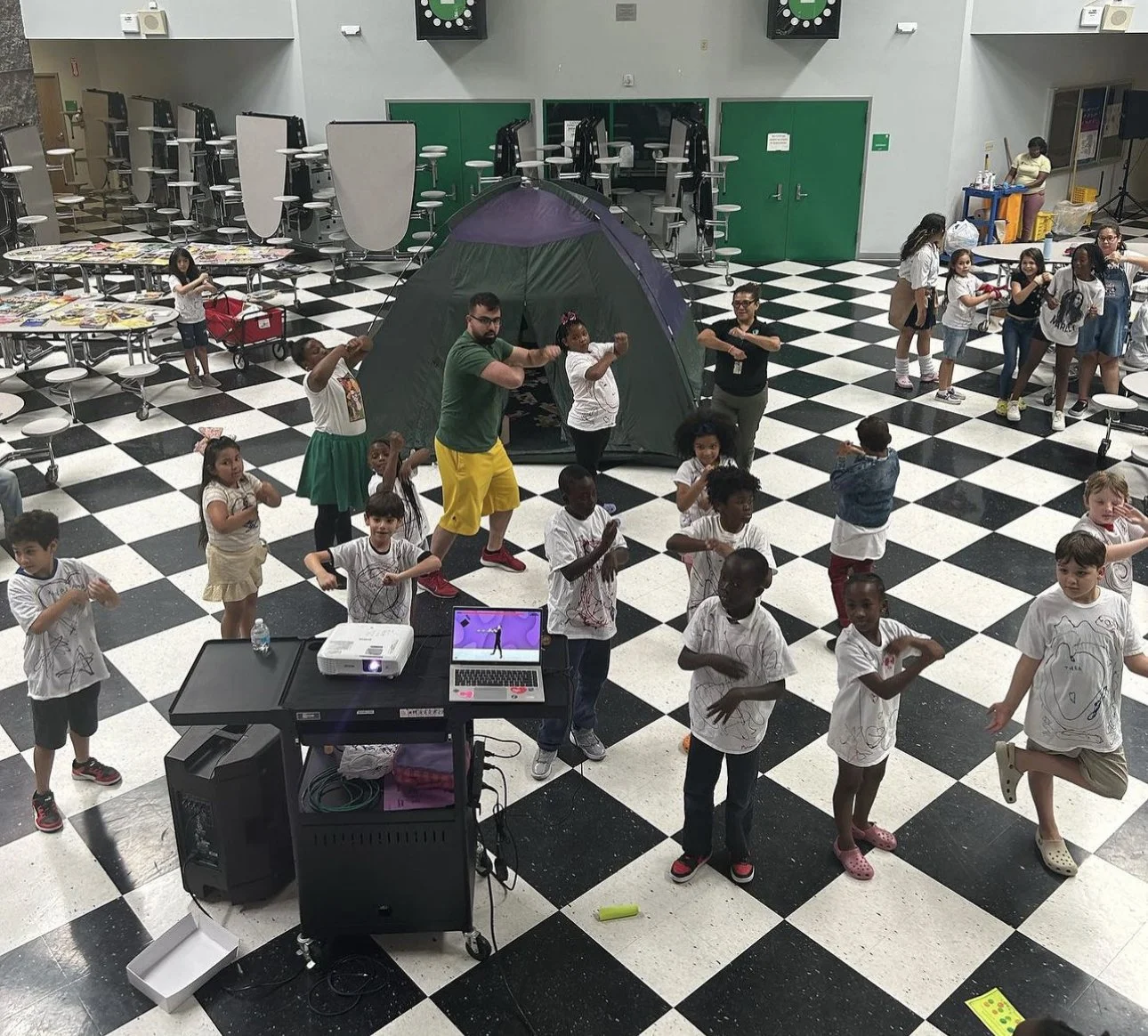 Children and adults participating in a dance or exercise activity in a large indoor space with a black and white checkered floor. A green tent is set up in the background, and there are tables and chairs stacked up on one side. A DJ setup with a lapt