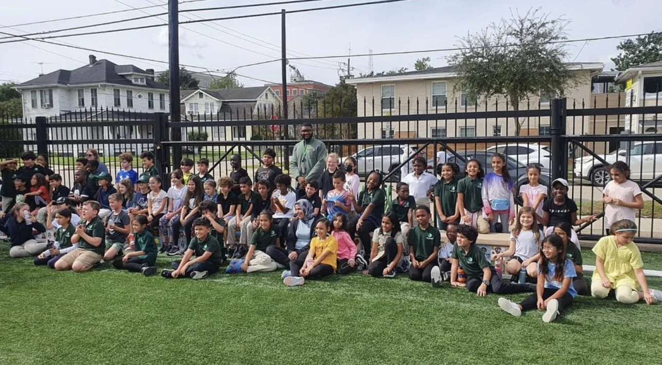 Group of children and two adults sitting and standing on a grassy field with a black fence and residential buildings in the background