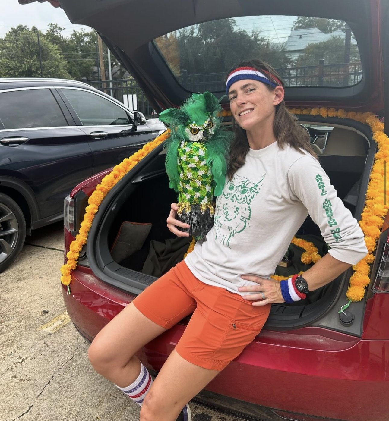 Man dressed in athletic clothing leaning on the open trunk of a red car outdoors, holding a colorful owl sculpture.