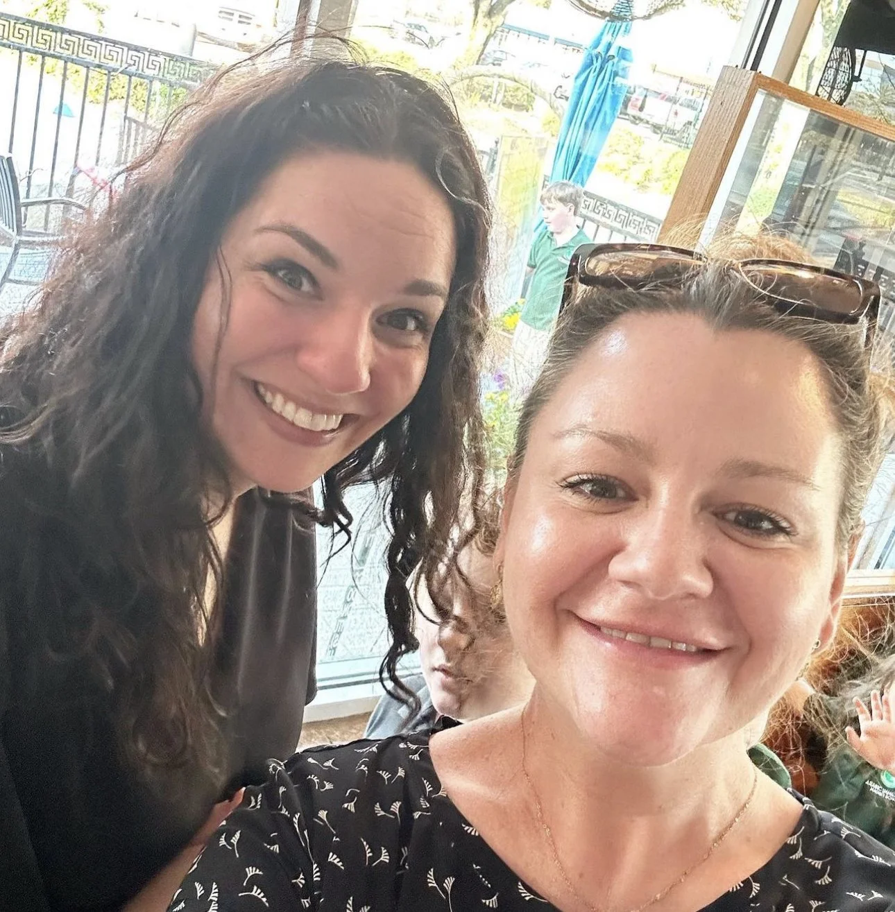 Two women smiling at a restaurant or cafe during daytime, with a window showing outdoor trees and a person walking outside in the background.