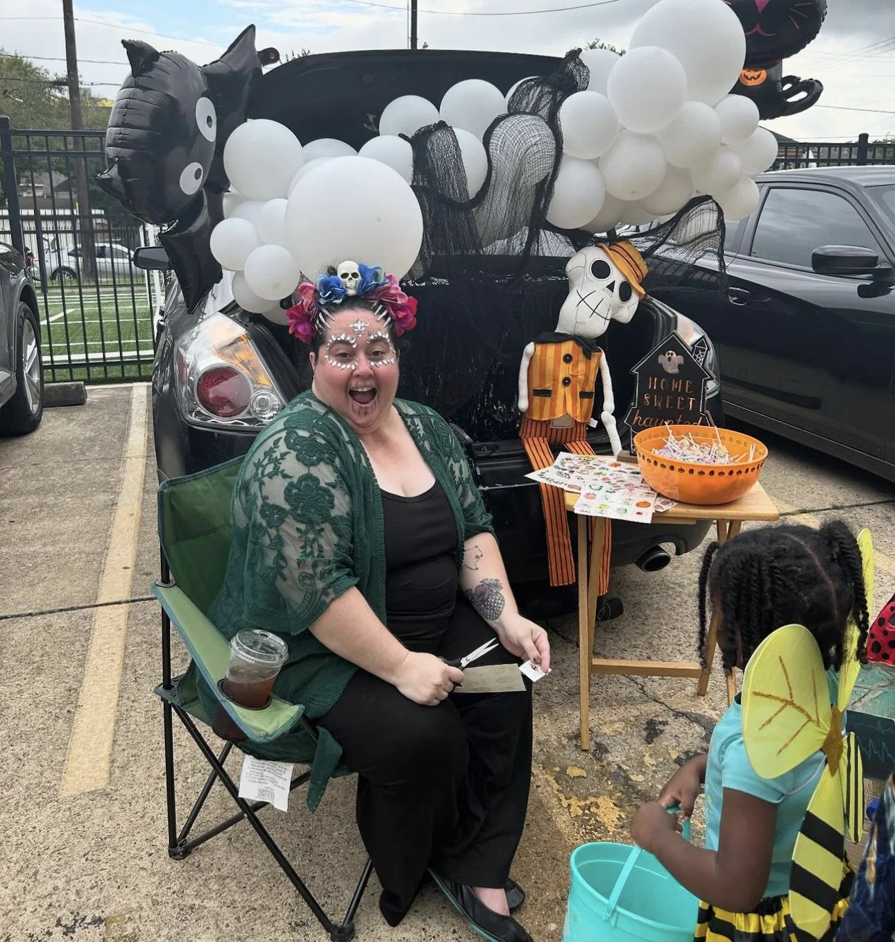 A woman with colorful face paint, wearing floral headband, sitting in a green camping chair, smiling and holding a knife, in front of a decorated car with Halloween themed balloons and decorations. A young girl with butterfly wings is nearby, holding
