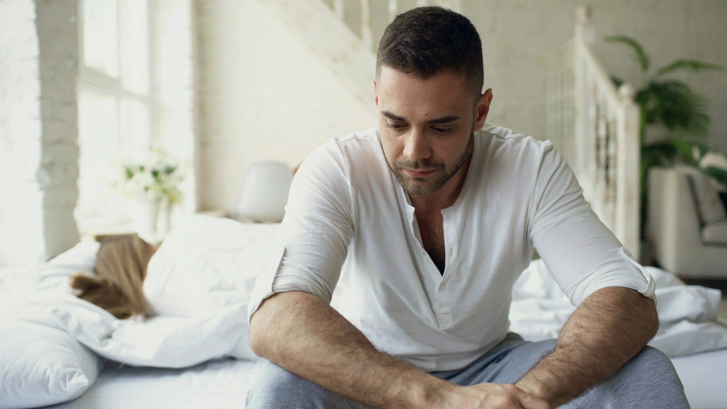 A man sits on a bed in a well-lit room, looking down thoughtfully. He has short dark hair, a beard, and wears a white shirt. The room has white brick walls, large windows, and houseplants.