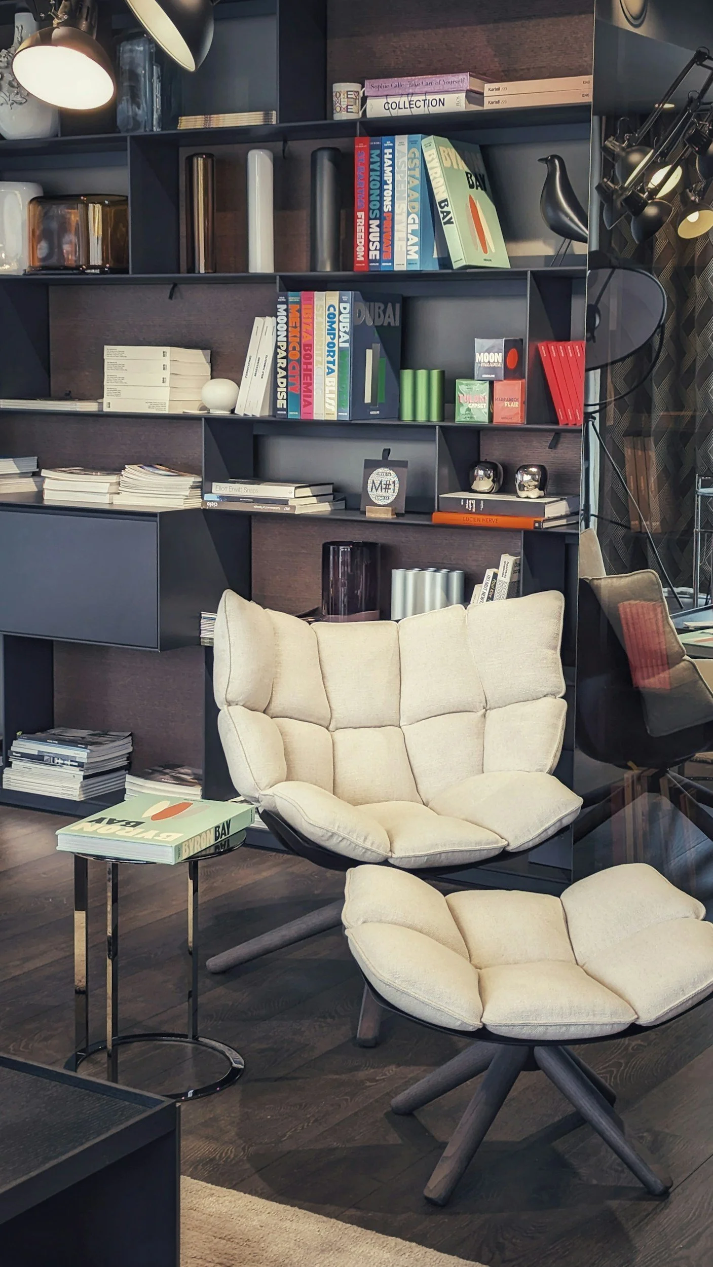 A cozy living space with two cream-colored cushioned chairs with wooden legs, a small side table with magazines, a dark bookshelf filled with books and decorative items, and additional chairs and lighting in the background.
