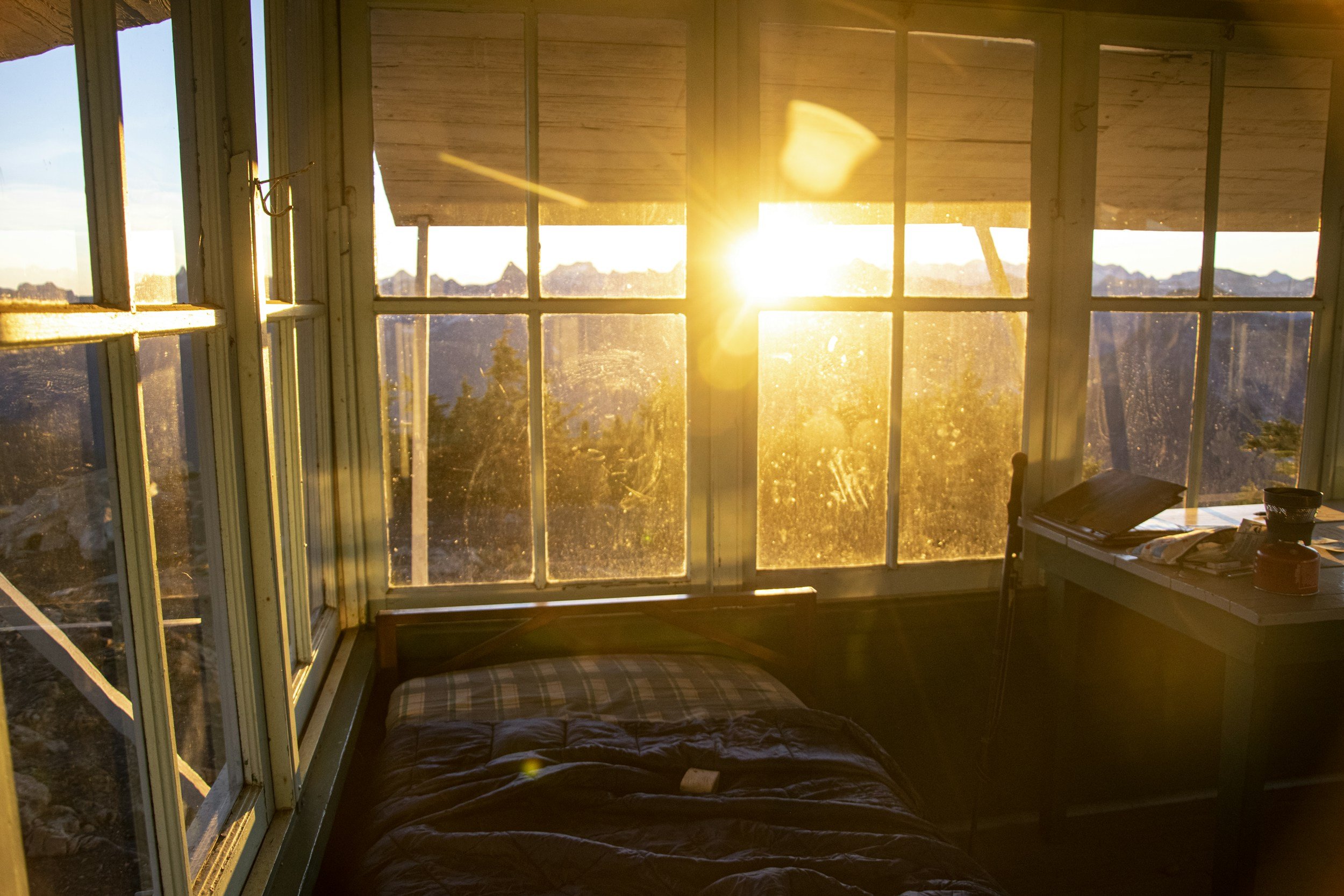 A cozy bedroom with large glass windows showing a mountain landscape during sunrise, a bed with a checkered pillow and dark bedding, and a small table with books and bowls.