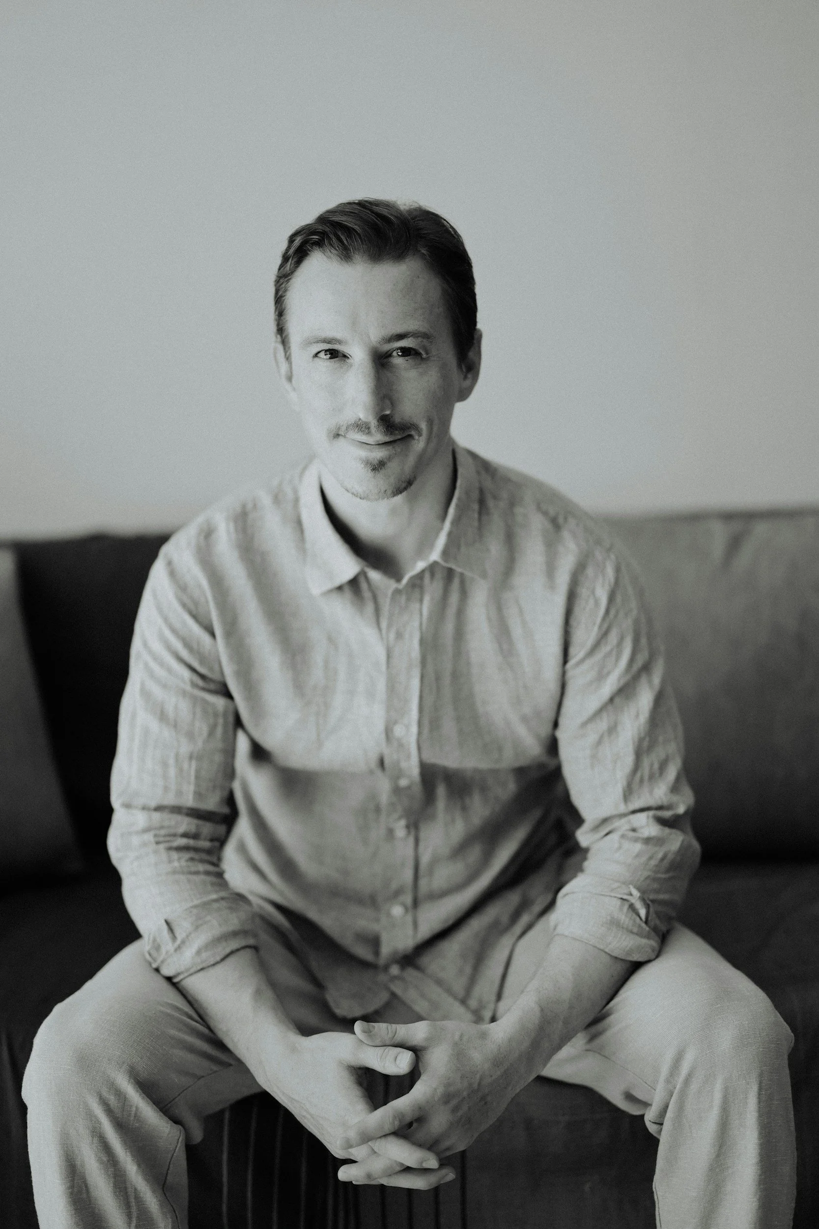 A black and white portrait of a man sitting on a dark sofa, looking at the camera with a slight smile, wearing a light-colored, long-sleeved, button-up shirt with the sleeves rolled up, and light-colored pants.