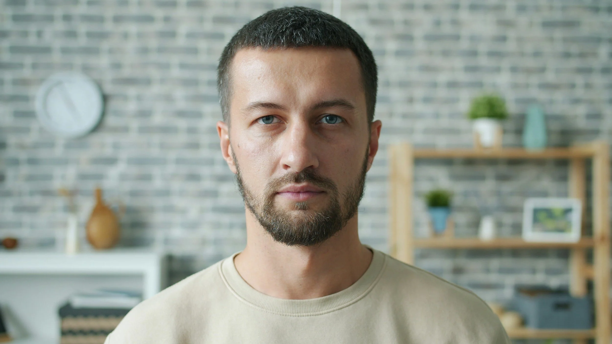 Close-up of a young man with blue eyes and a beard, standing indoors with a brick wall background and shelves with decorative items.