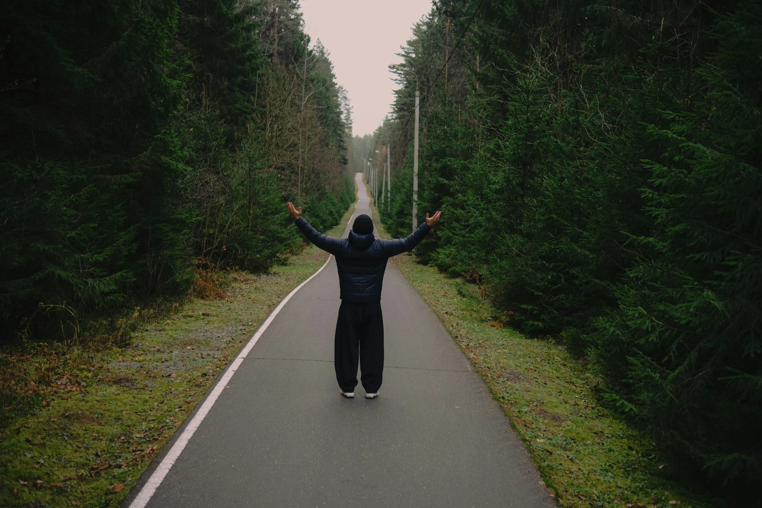 A person standing on a quiet, paved road in the woods with arms raised, surrounded by green trees on both sides.