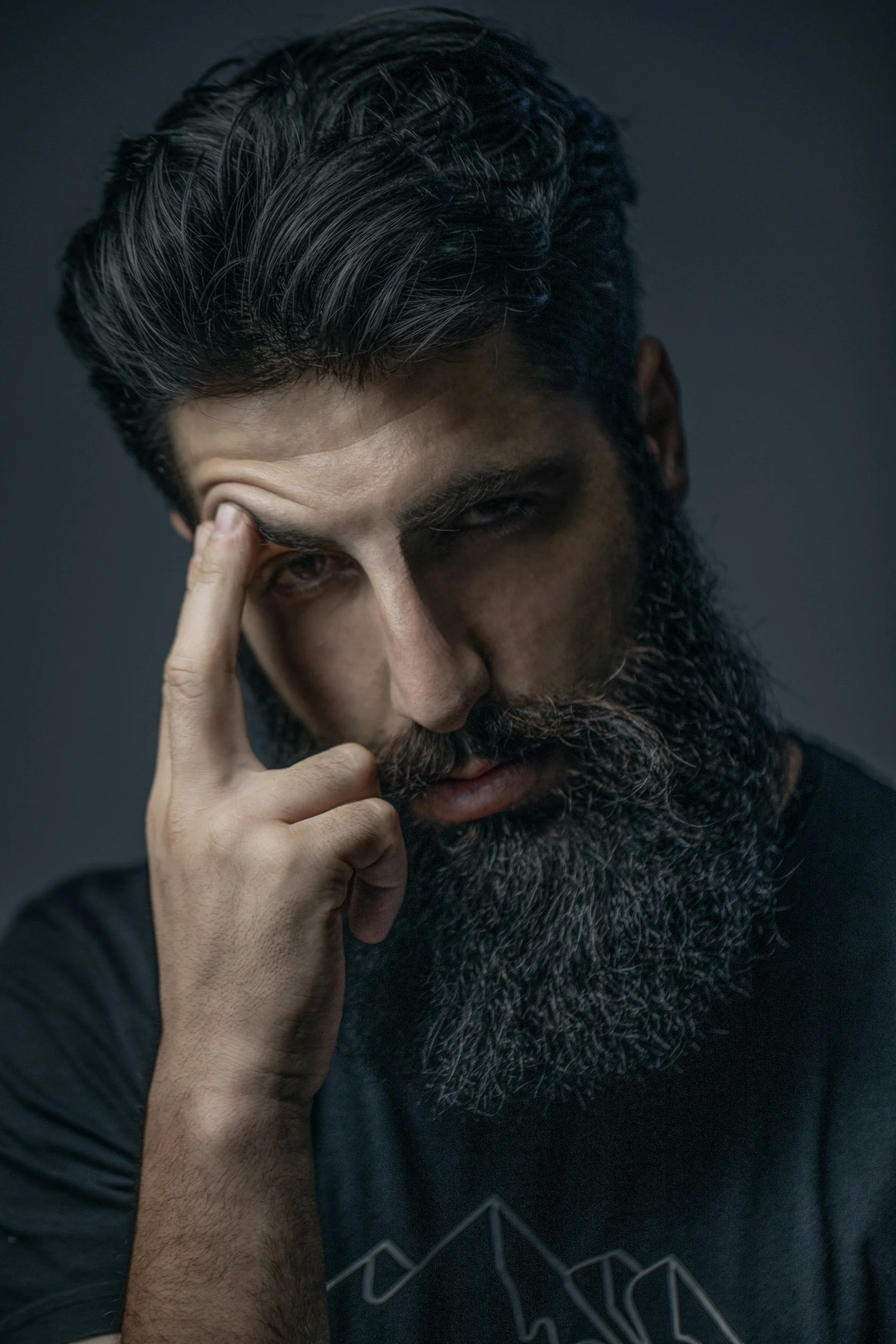 A man with dark hair and a full beard, touching his forehead with his fingers, looking intently at the camera with a serious expression.