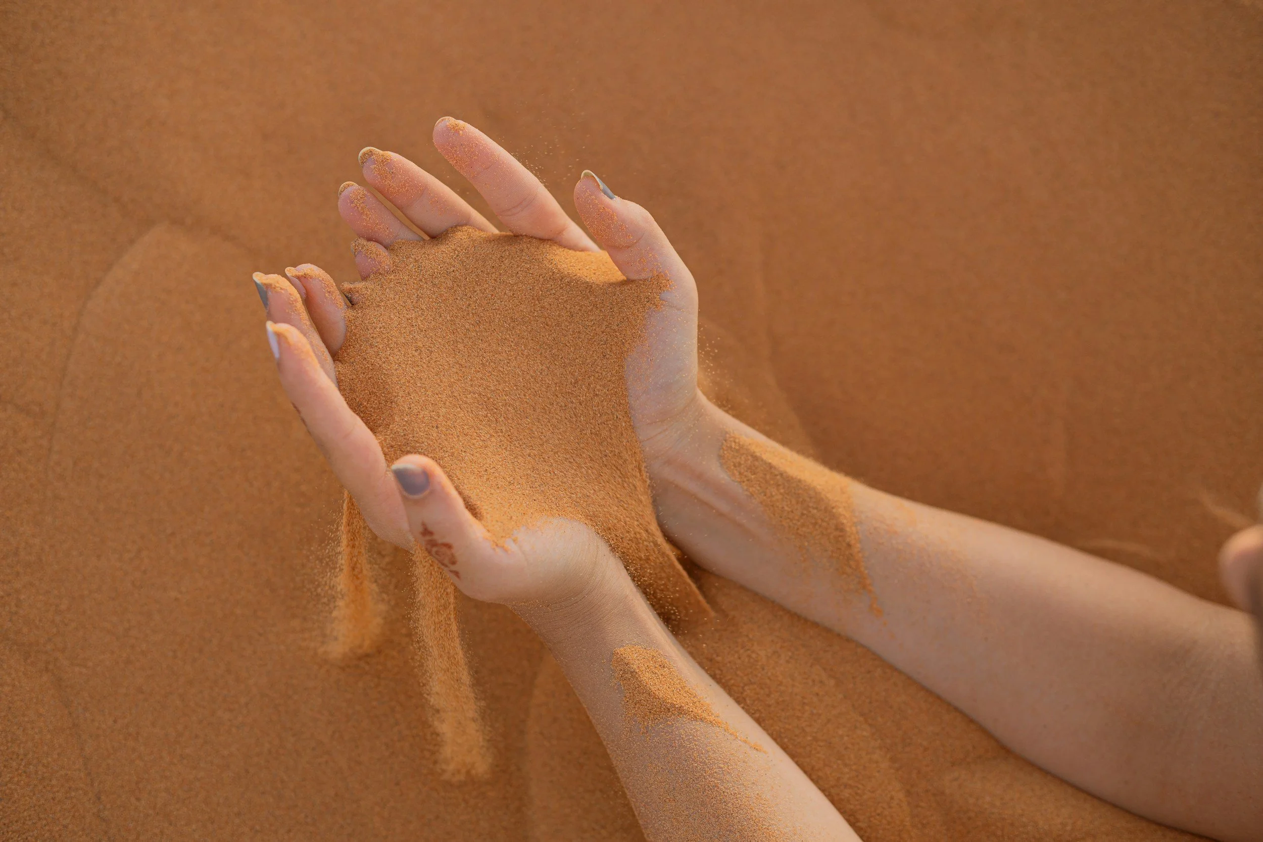 Hands holding and pouring fine orange-brown sand, creating a small pile on a sandy surface.