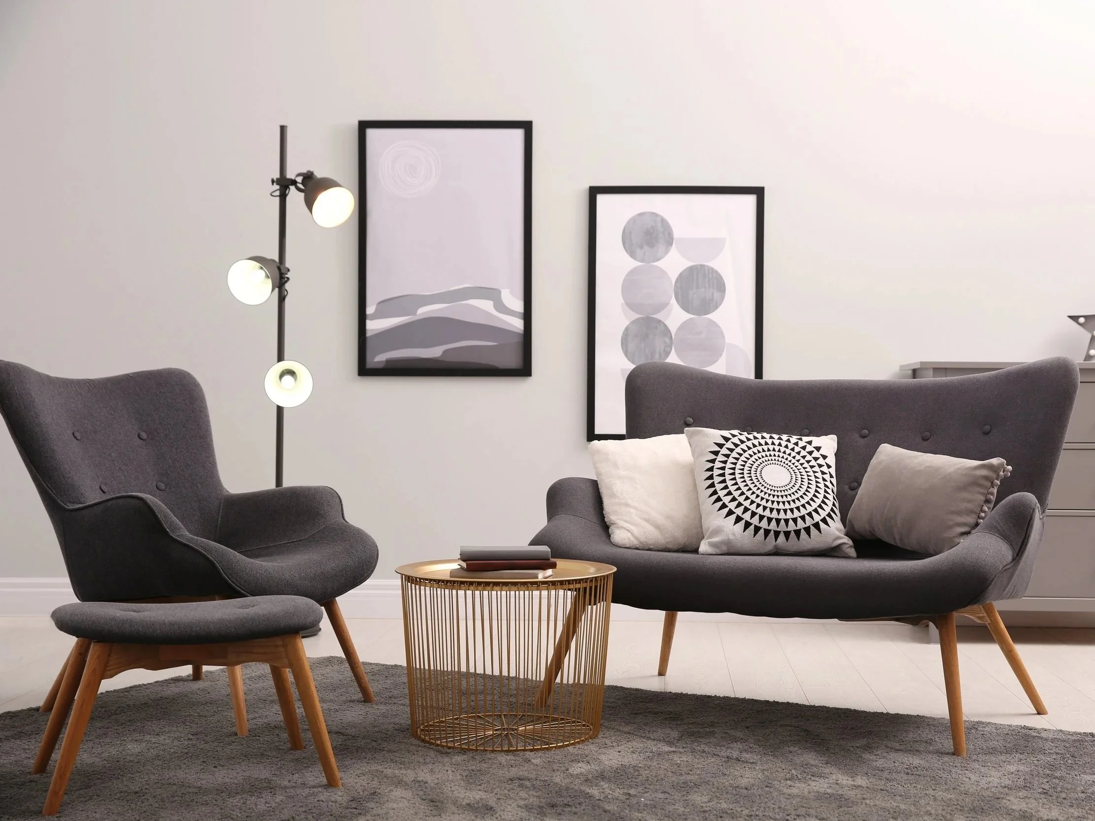 Modern living room with gray armchair, sofa with decorative pillows, gold wire coffee table, black floor lamp with three lights, and abstract black and white artwork on white walls.