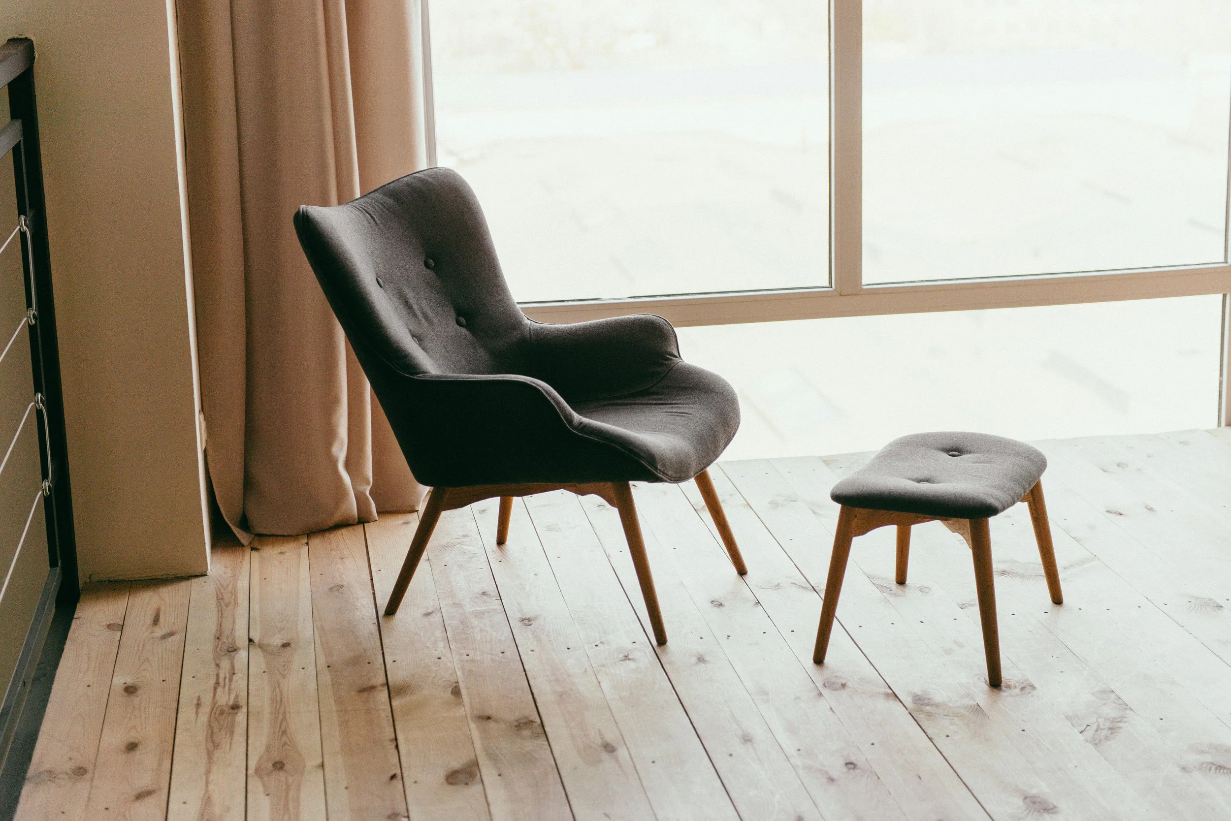 A cozy corner with a black upholstered armchair and matching ottoman by a large window, with beige curtains and light wooden flooring.