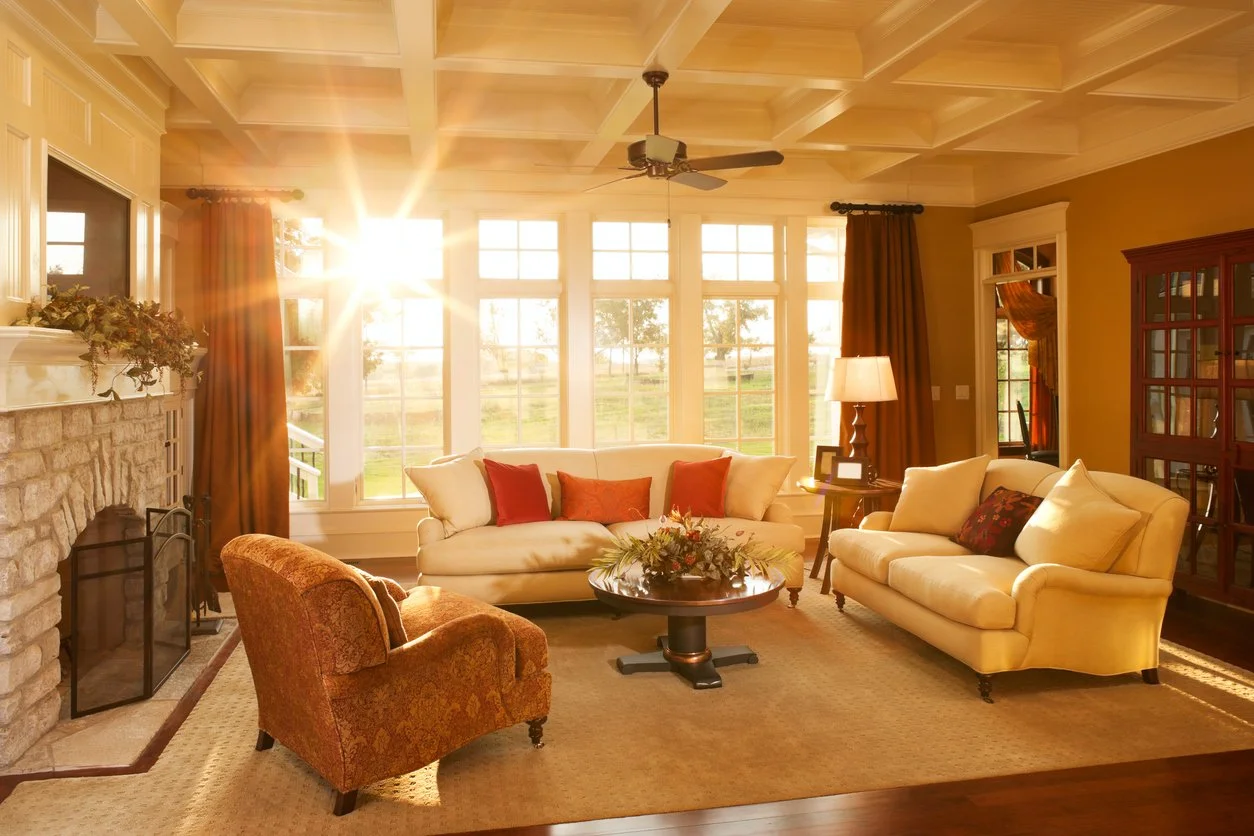 Living room with large windows, sunlight streaming in, cream sofas, a patterned armchair, a fireplace with greenery, a ceiling fan, a coffee table with a floral centerpiece, and warm-toned curtains and walls.