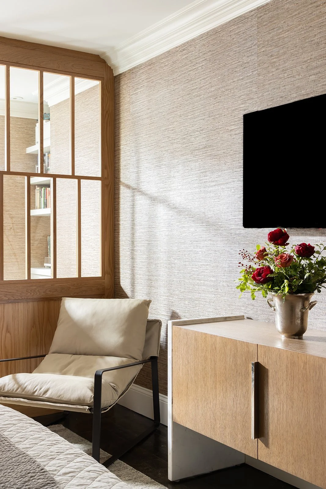 Cozy loft bedroom reading nook featuring a modern accent chair, textured beige wallpaper, a light brown wood room divider screen, and a custom media console styled with colorful flowers on a tan rug.