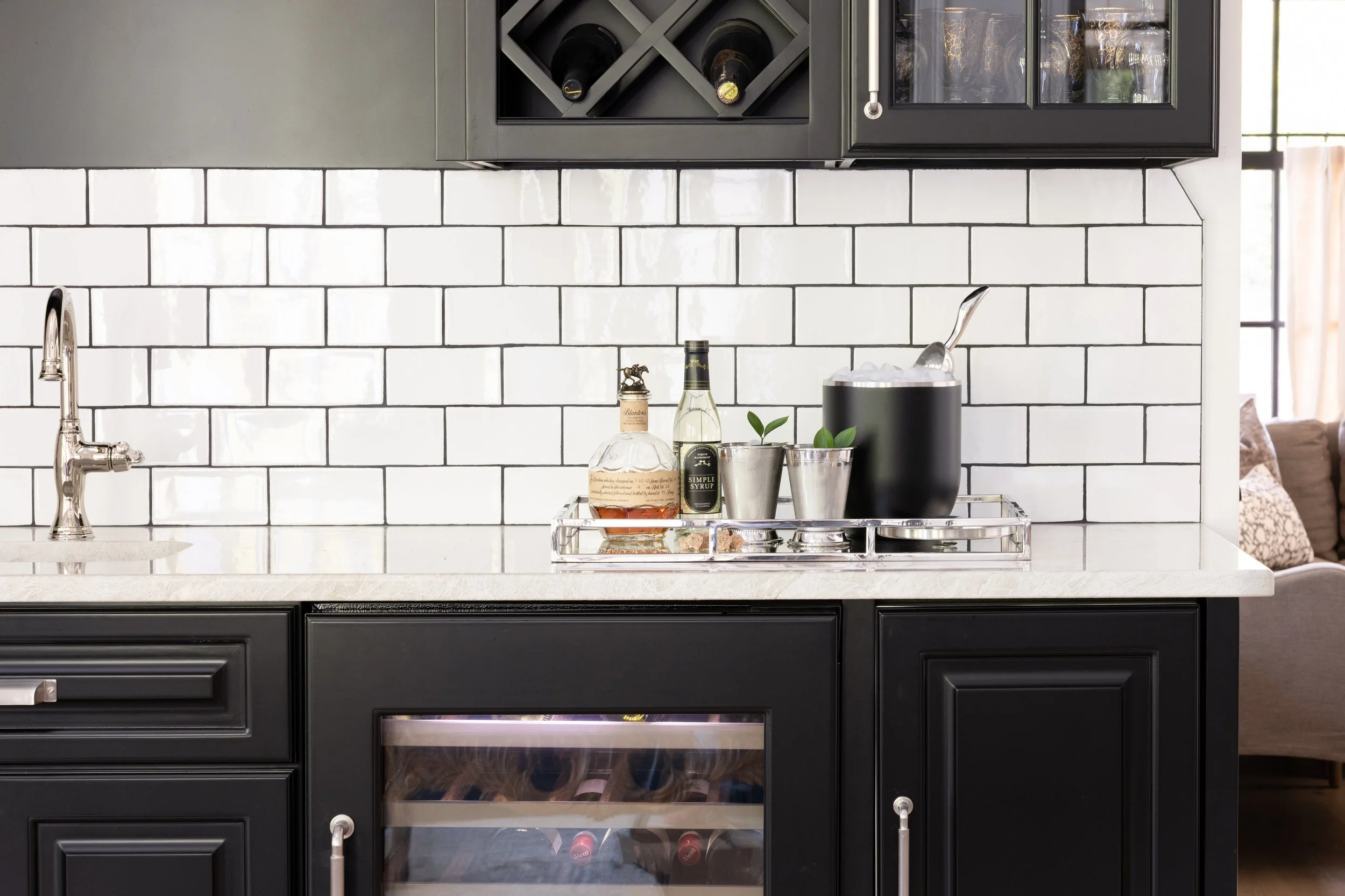A kitchen countertop with a black wine rack, a cabinet with glasses, and various bottles and plants on a tray.